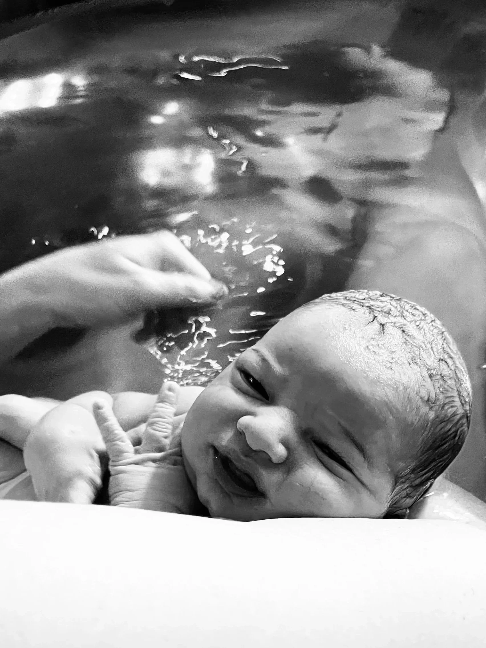 A newborn baby being bathed in water, with a hand gently supporting the baby's head. The baby has wet hair and is smiling.