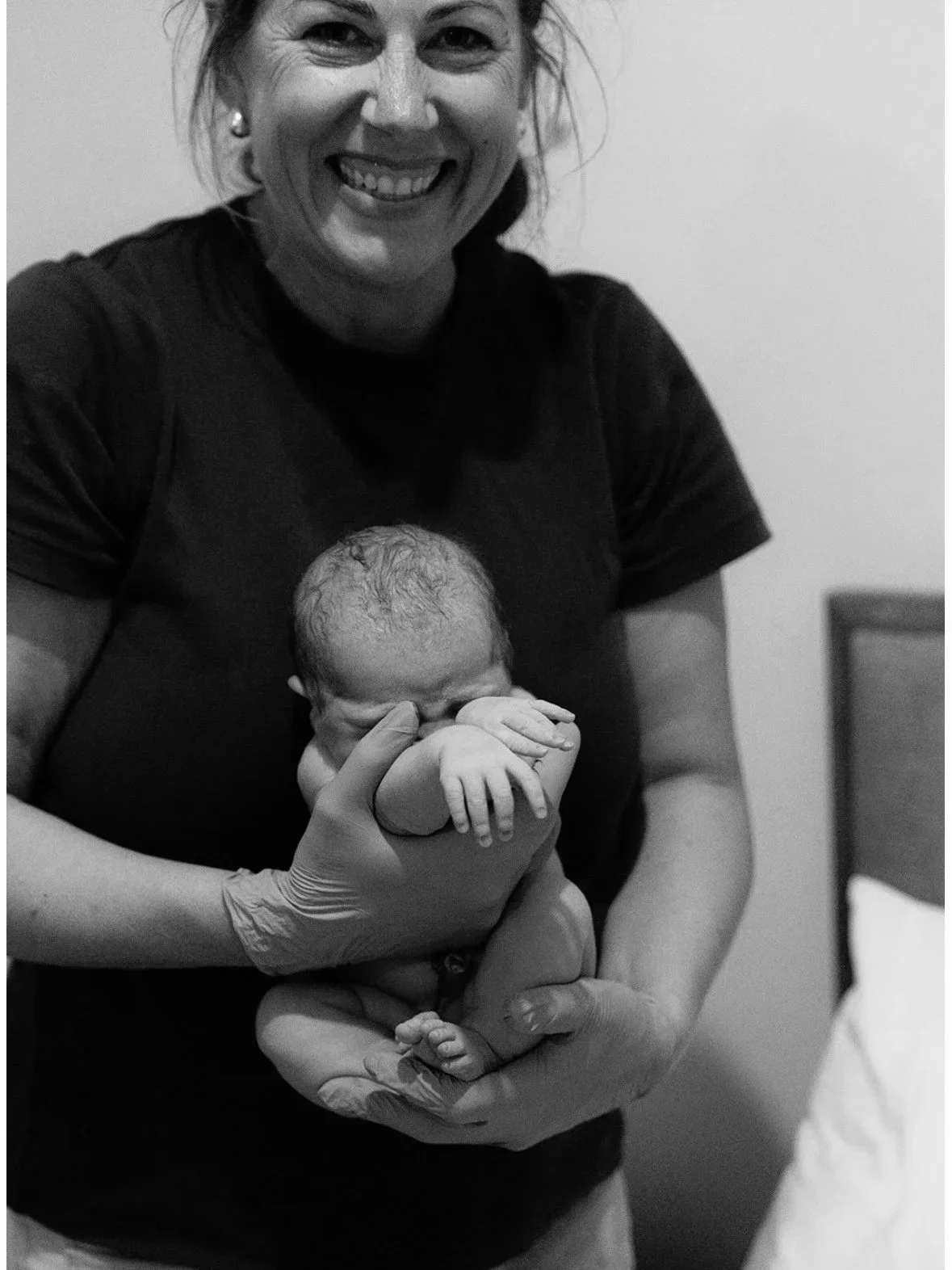 A woman smiling and holding a newborn baby, with the woman wearing gloves.