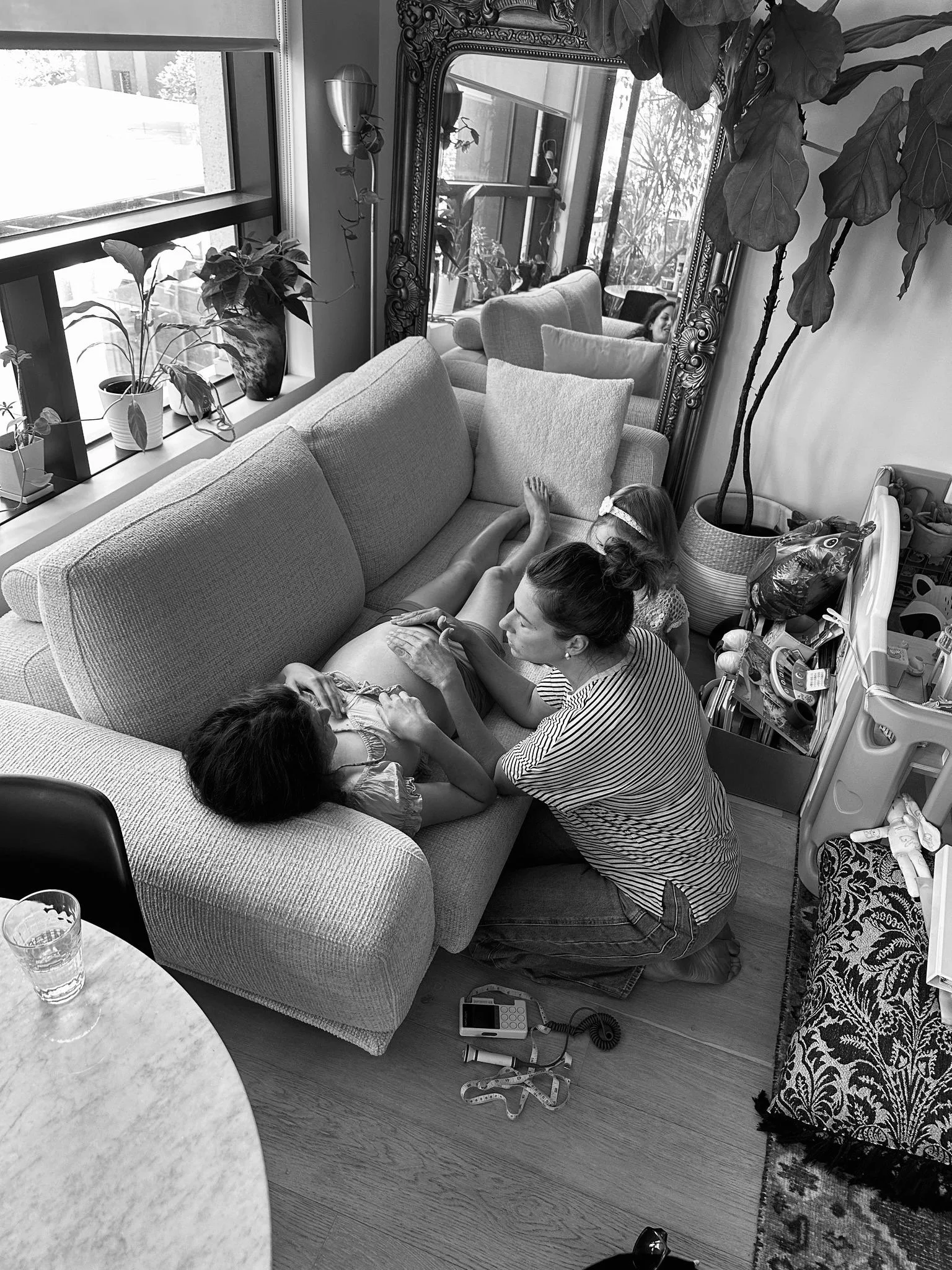 A woman and two children on a couch with a pregnant woman inside a cozy living room, with plants on the window sill and a large ornate mirror on the wall.