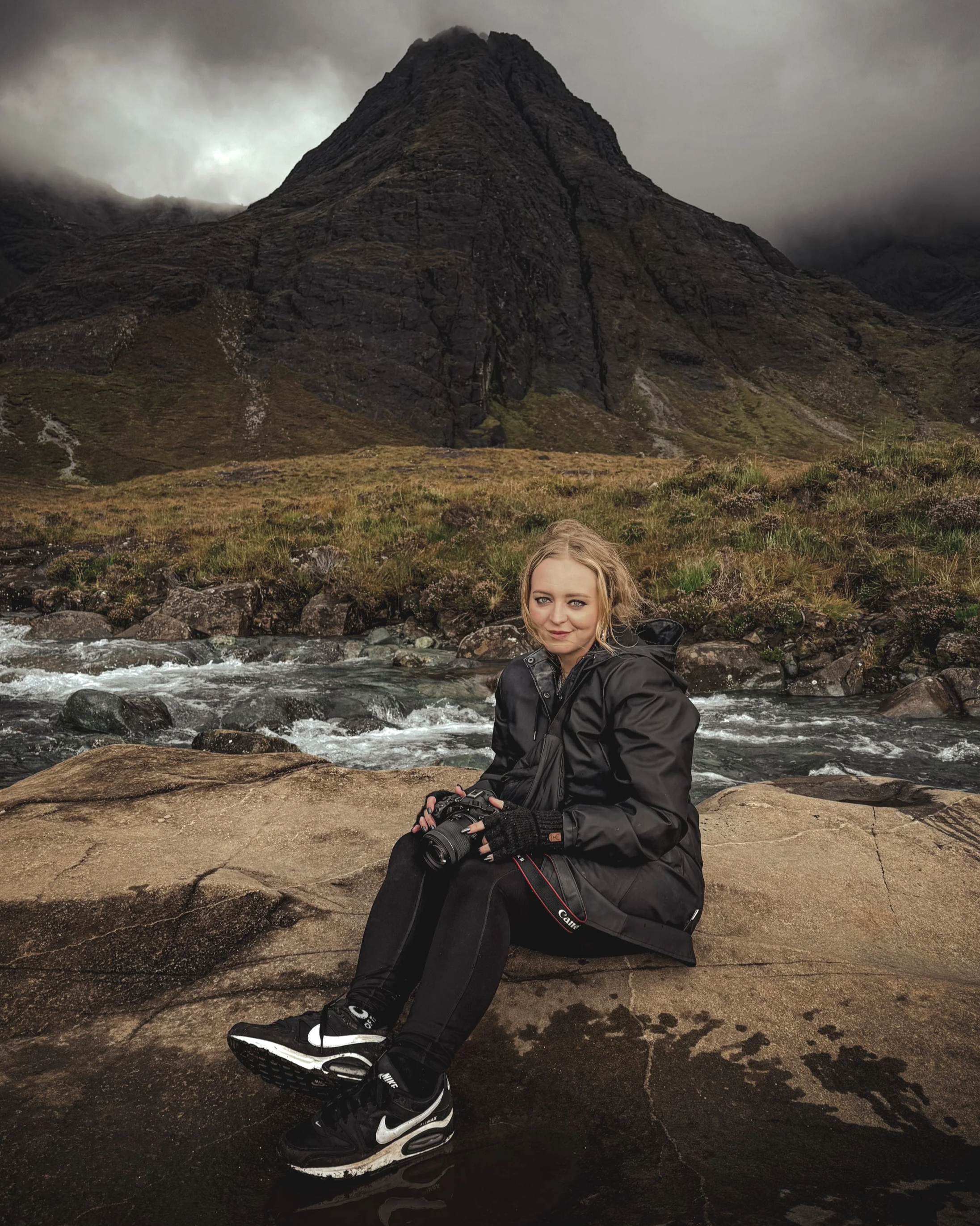 A woman with blonde hair and blue eyes sitting on a rocks by a stream with a mountain in the background, wearing a black jacket, black pants, black gloves, and holding a camera.