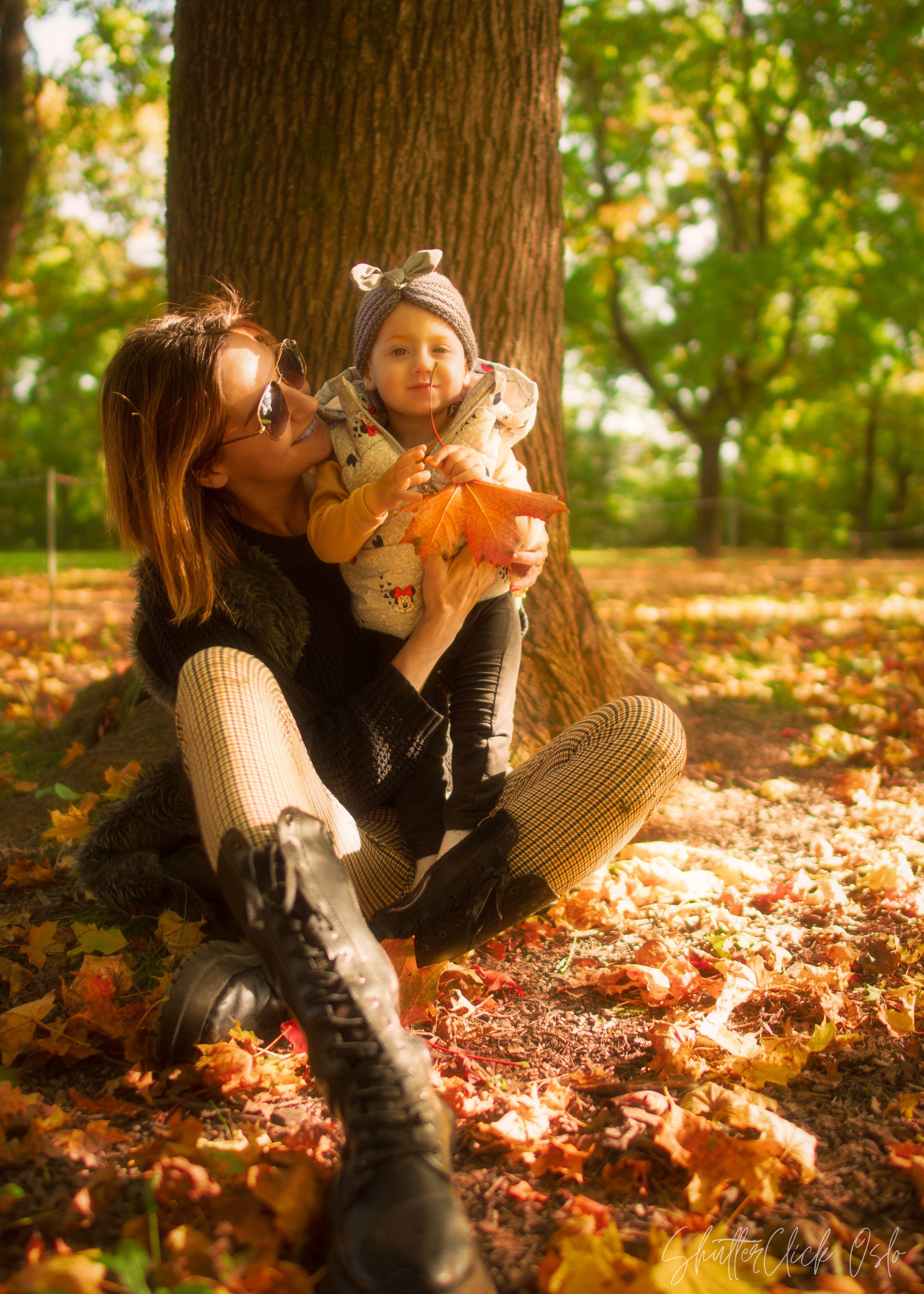 A woman sitting on the ground in a park holding a toddler, both enjoying autumn leaves. The child is holding a large orange leaf, and both are dressed warmly with a tree trunk behind them. The ground is covered with fallen leaves.
