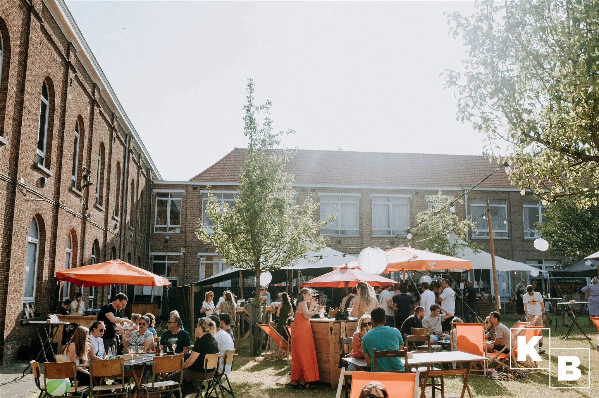 Een drukke feestlocatie in een binnenplaats met mensen die aan tafels zitten en praten, er staan parasols en lampionnen hangen aan draden tussen de bomen. Op de achtergrond een gebouw met ramen en een daken.