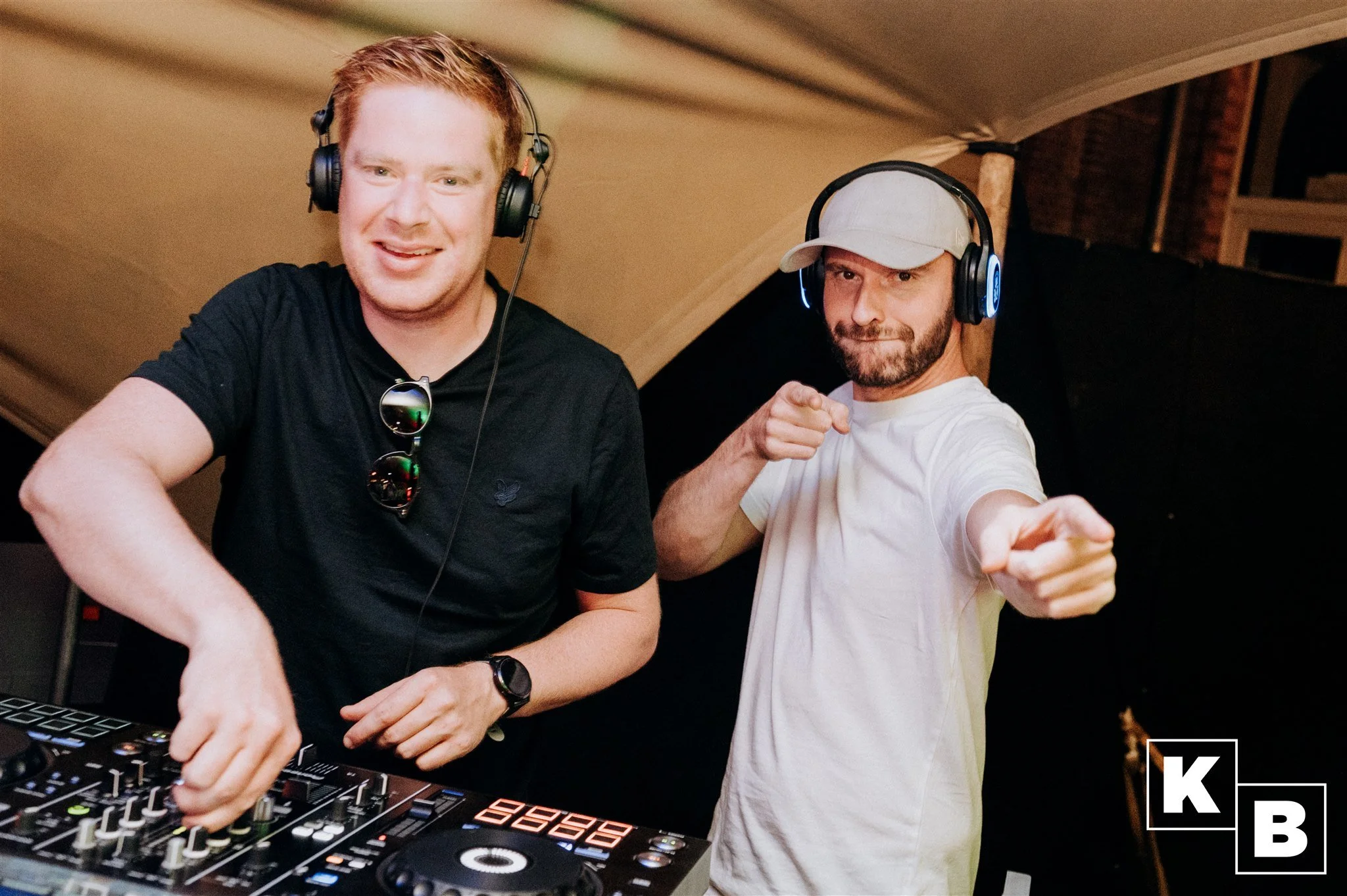 Twee mannen met koptelefoons die samen draaien in een club of studio, de ene wijst naar de camera, de andere draait op DJ-apparatuur.