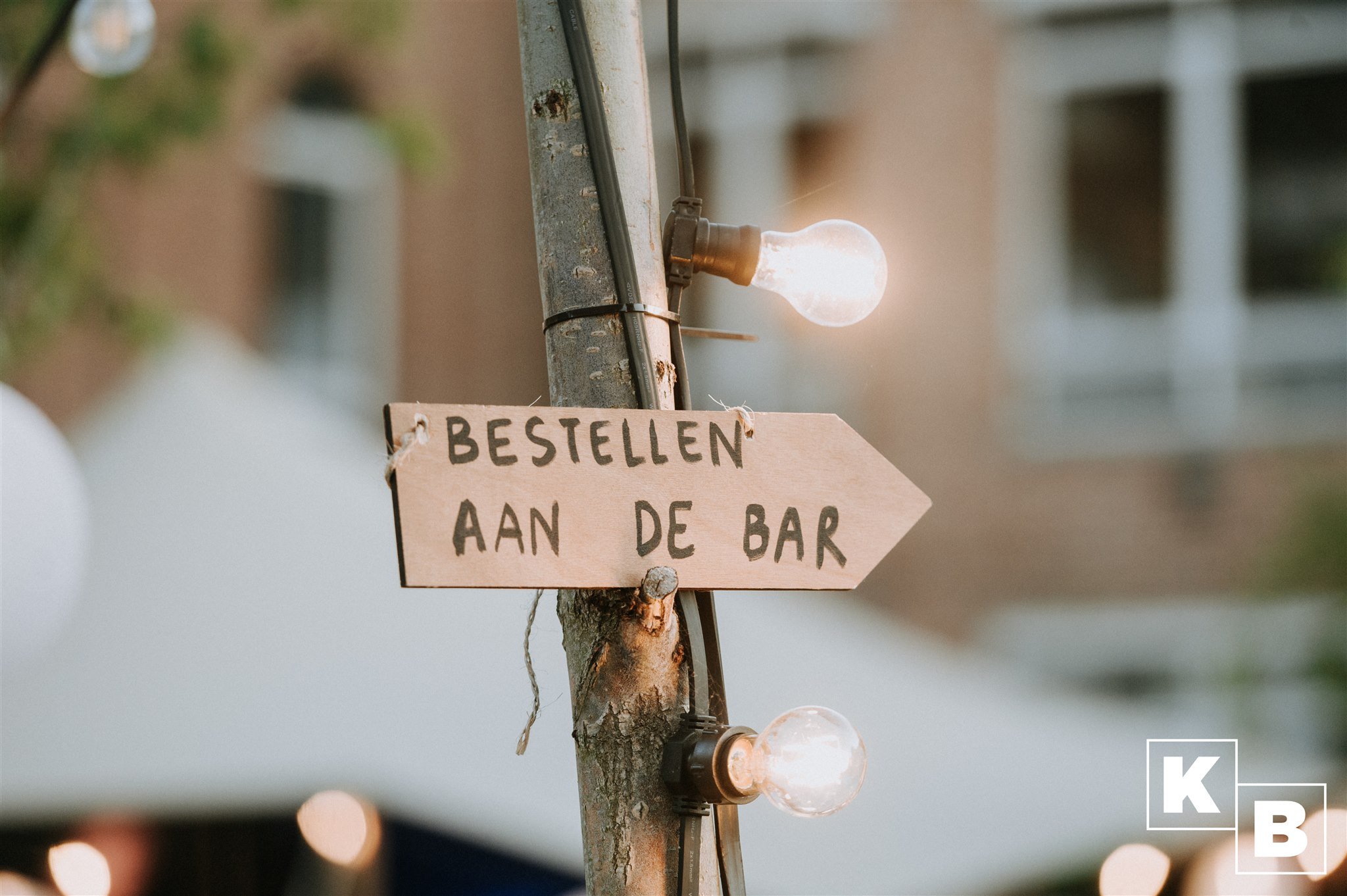 Een houten bord aan een boom met de tekst 'Bestellen aan de bar', verlicht door een sfeervolle gloeilamp.