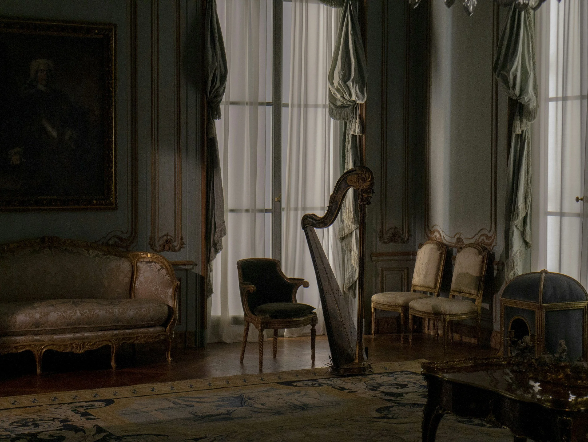 An ornate classical living room with antique furniture, including a sofa, chairs, a harp, and a decorative chest, with large windows covered by elegant curtains.