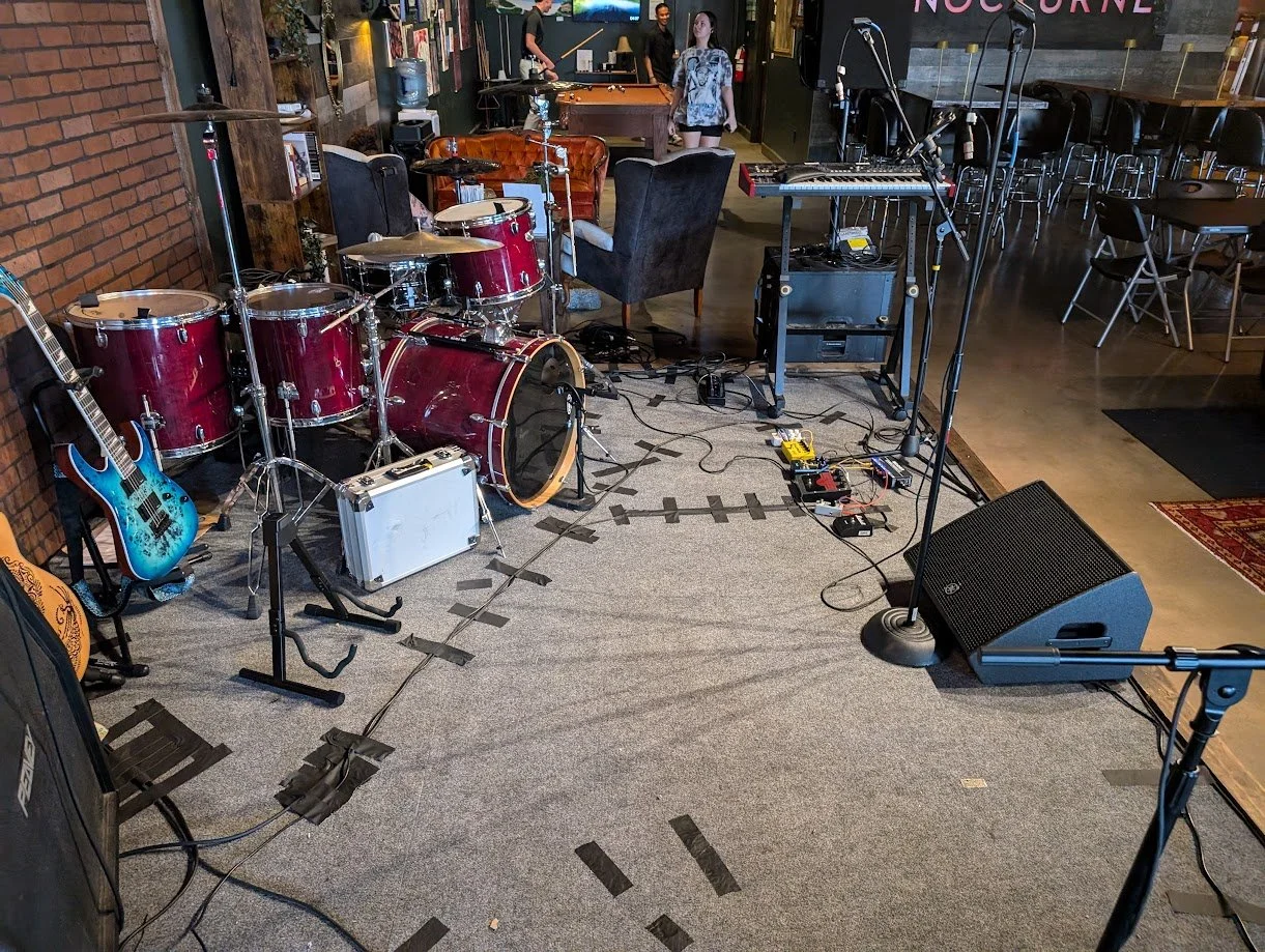 A stage with a drumset, guitar, guitar amplfier, floor monitor, and microphone visible with cables taped to the floor