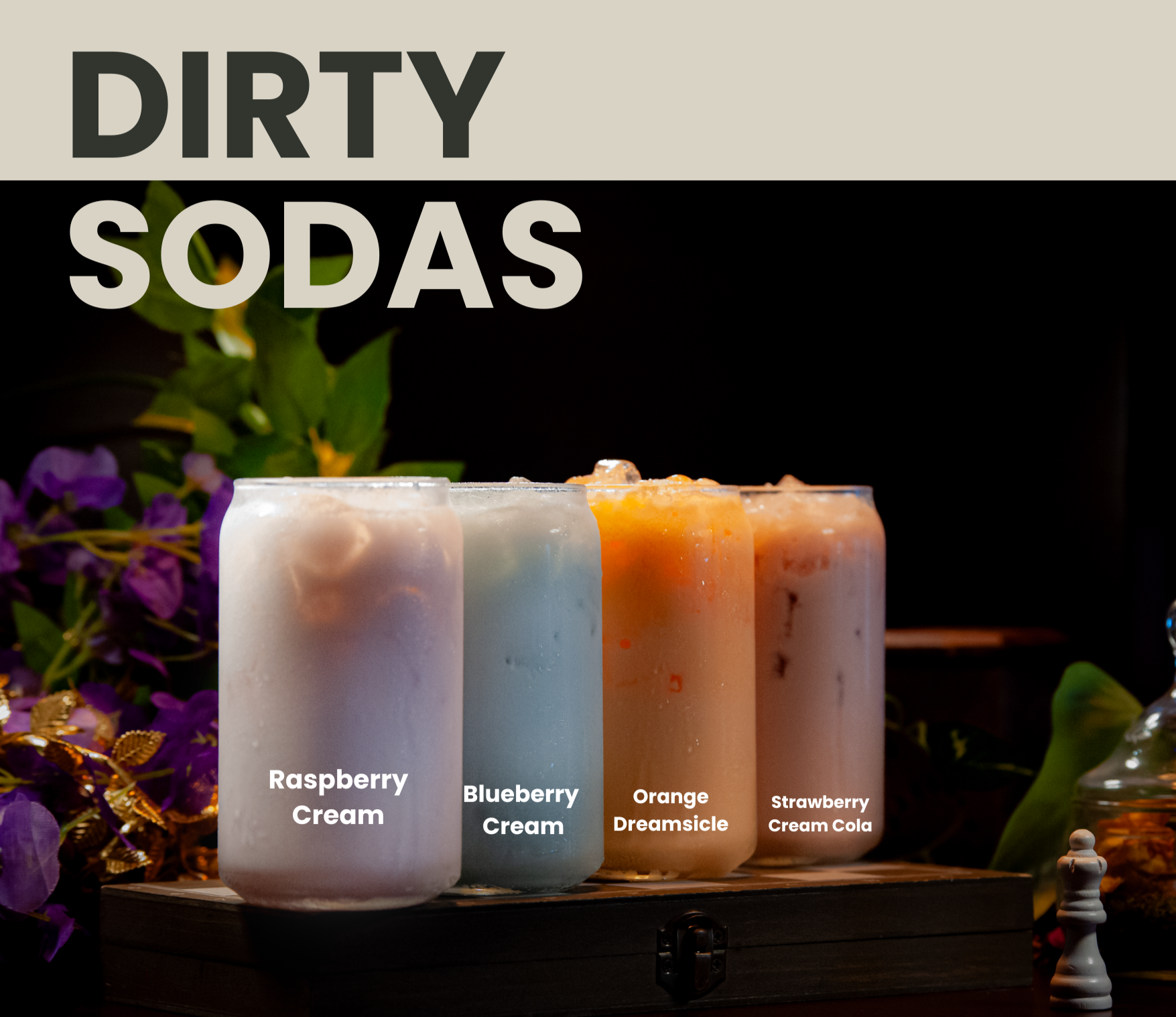 Three cups containing dirty sodas sitting on top of a raised platform with foliage in the background. The cups contain ice, the liquids are solid in color