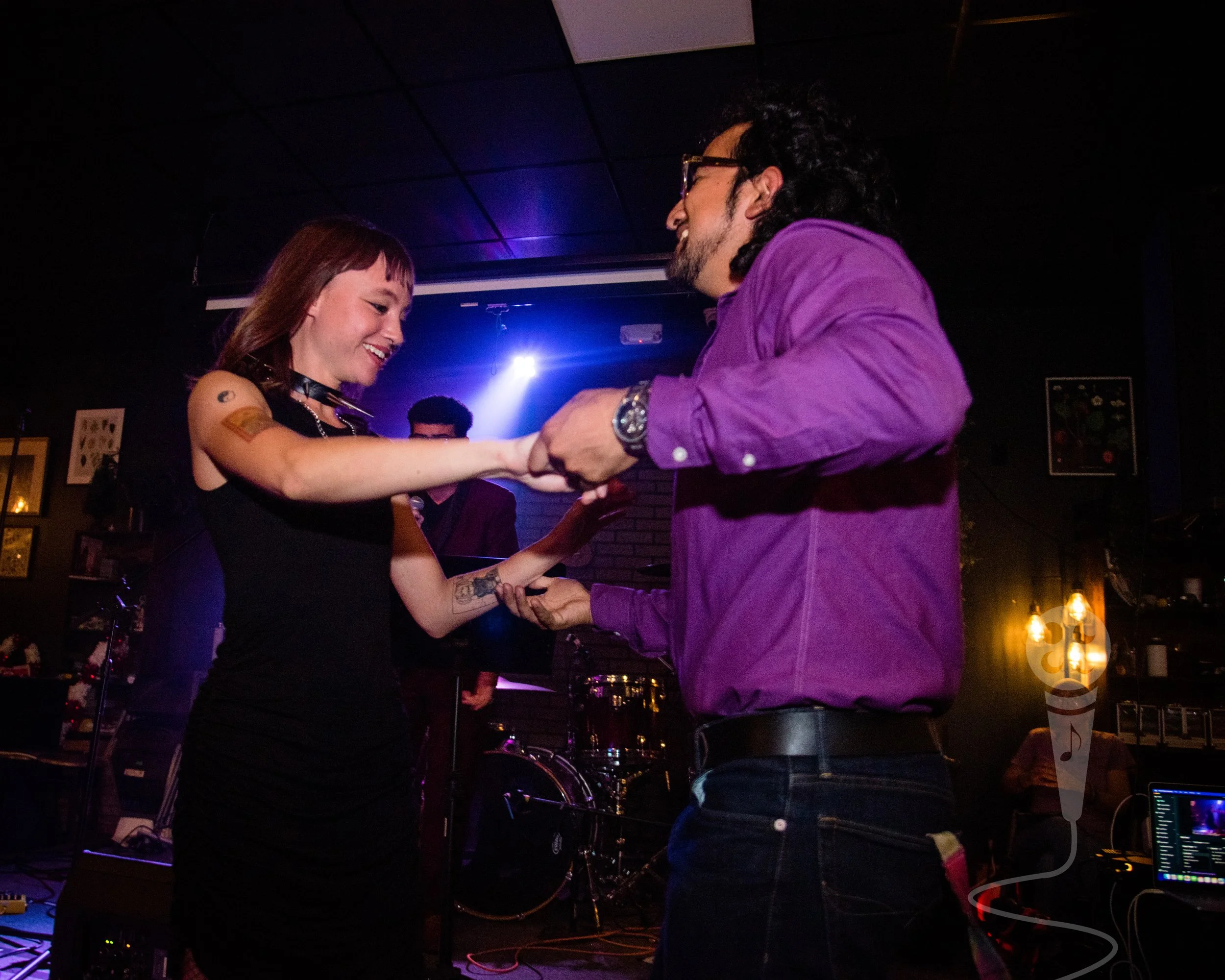 Two individuals holding hands and dancing. There is a stage with performers in the background with a spotlight shining on the lead singer.