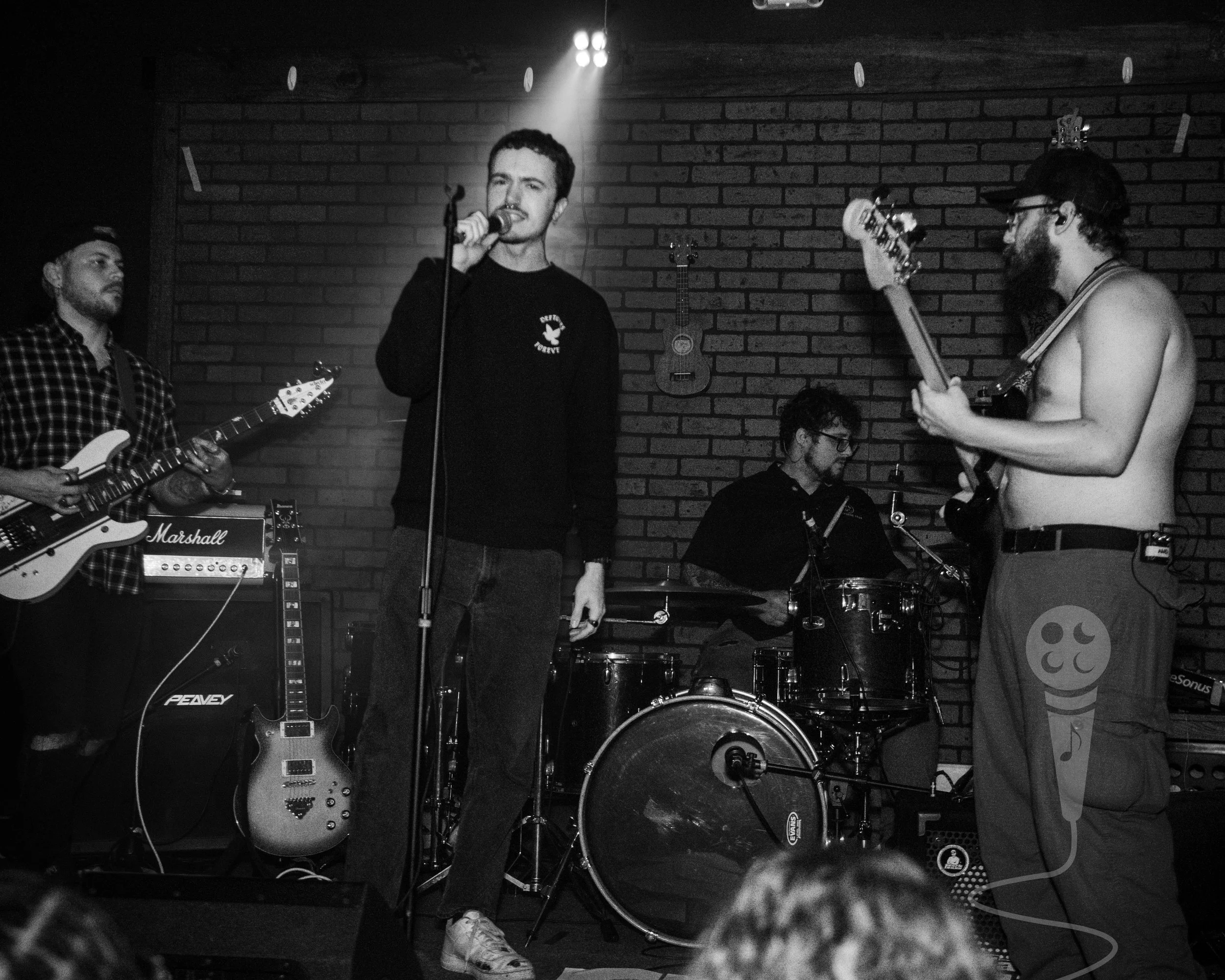 A black and white image of a band performing on stage. Singer is center stage with a microphone in right hand and spotlight shining on his back. Bass player is on the right, shirtless, the guitar player on the left, drummer in background.