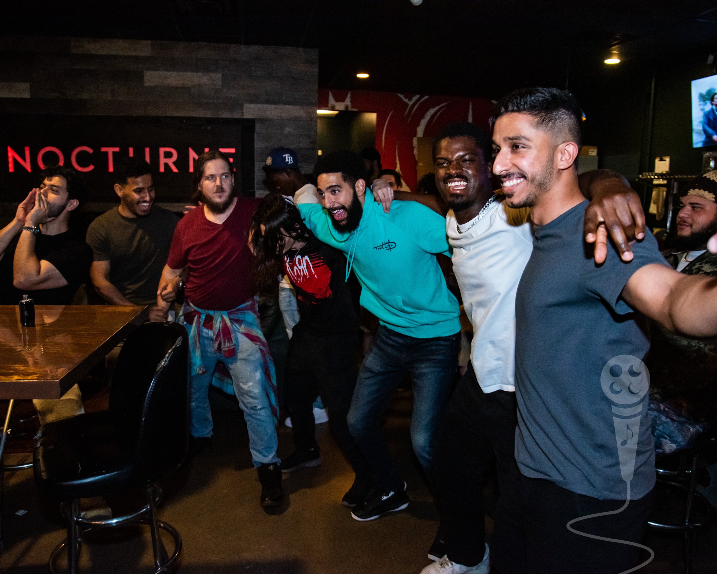 Three men standing with their arms on each others shoulders smiling, with other males and females in the background, all look happy and appear to be having a good time