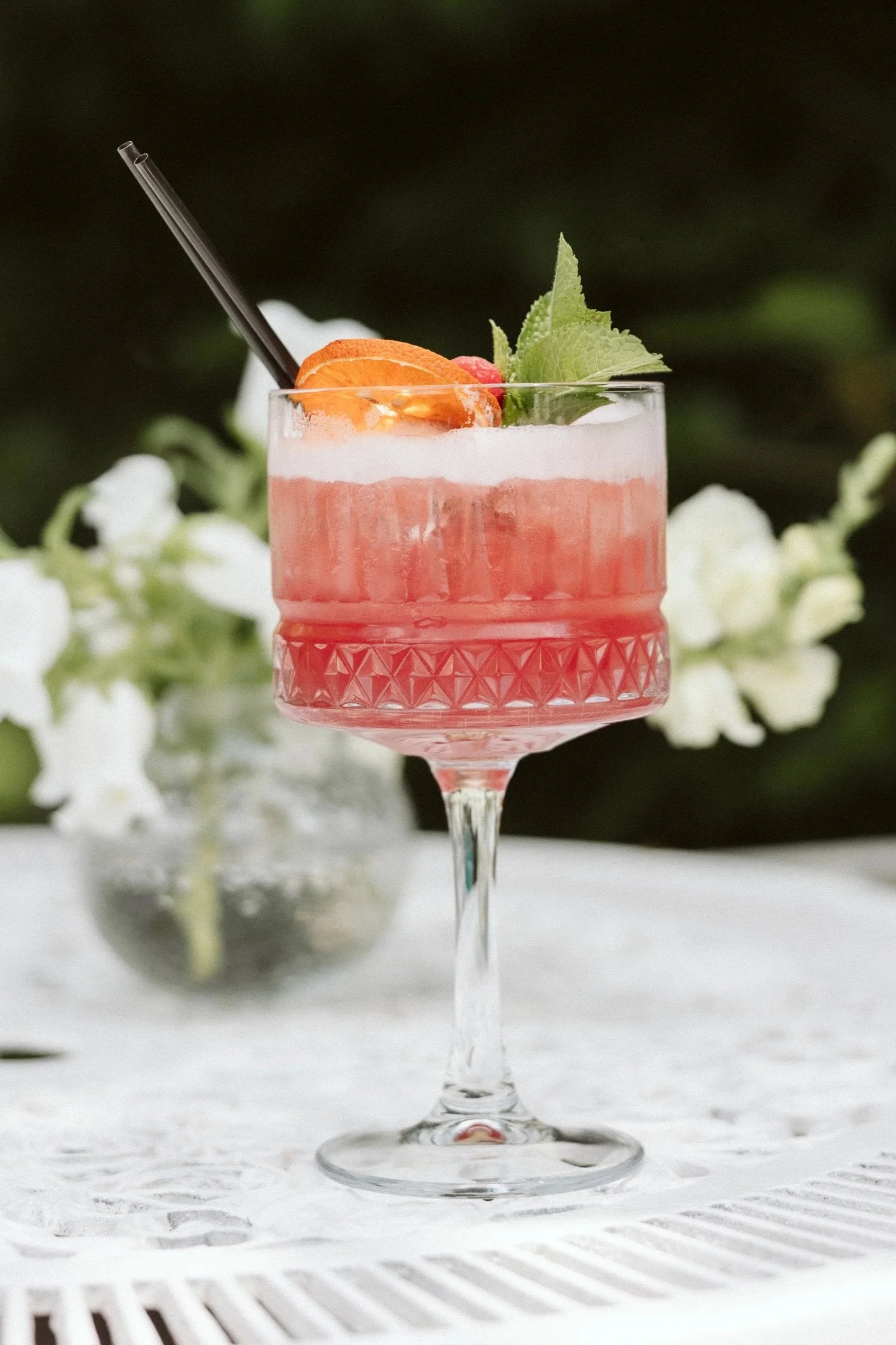 A pink cocktail in an elegant glass garnished with orange slices, mint leaves, and a raspberry, on a white table with a blurred background of white flowers.