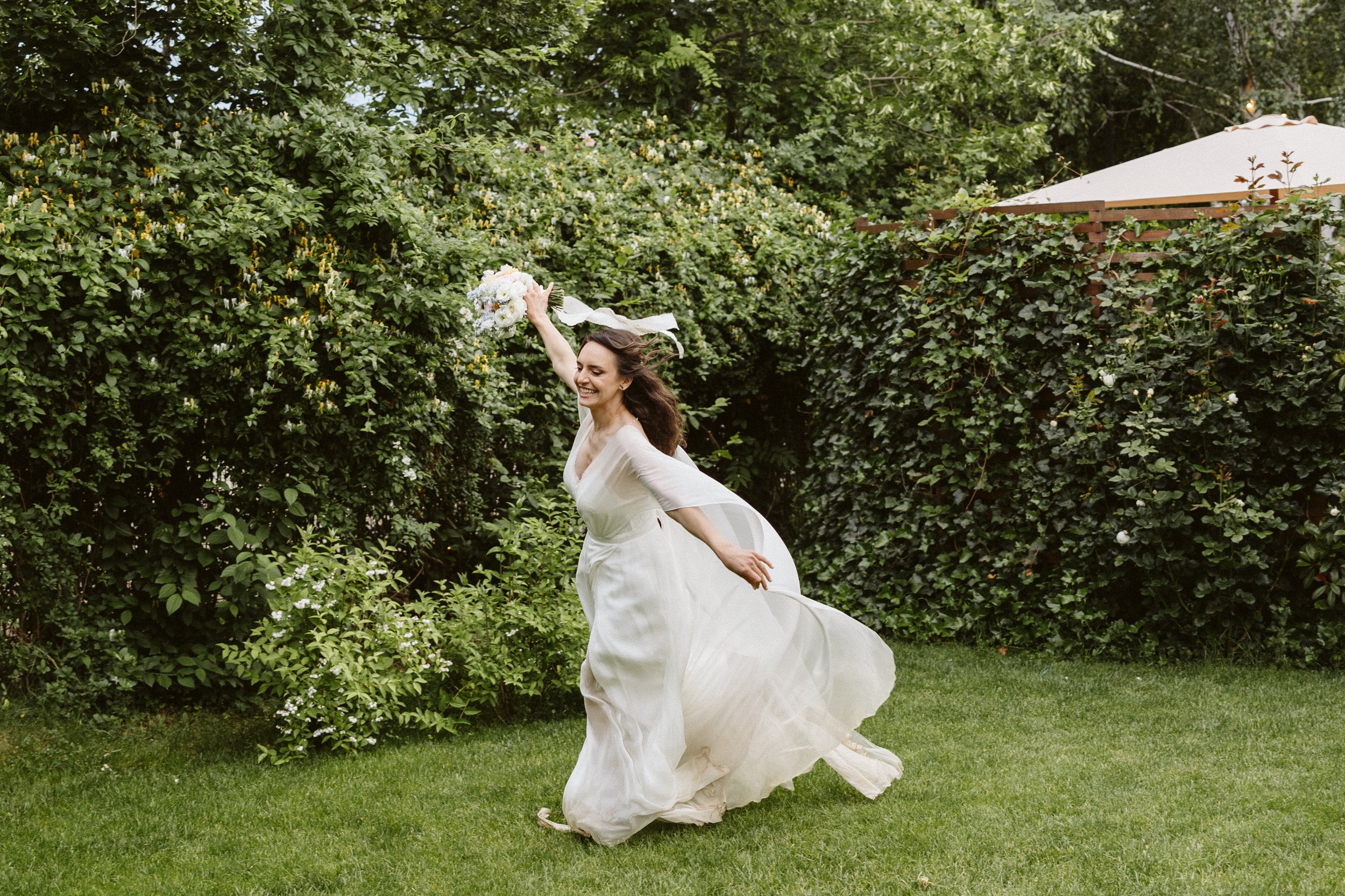 A woman in a wedding dress dancing joyfully on a green lawn, holding a bouquet of flowers, with a background of lush greenery and a white umbrella.