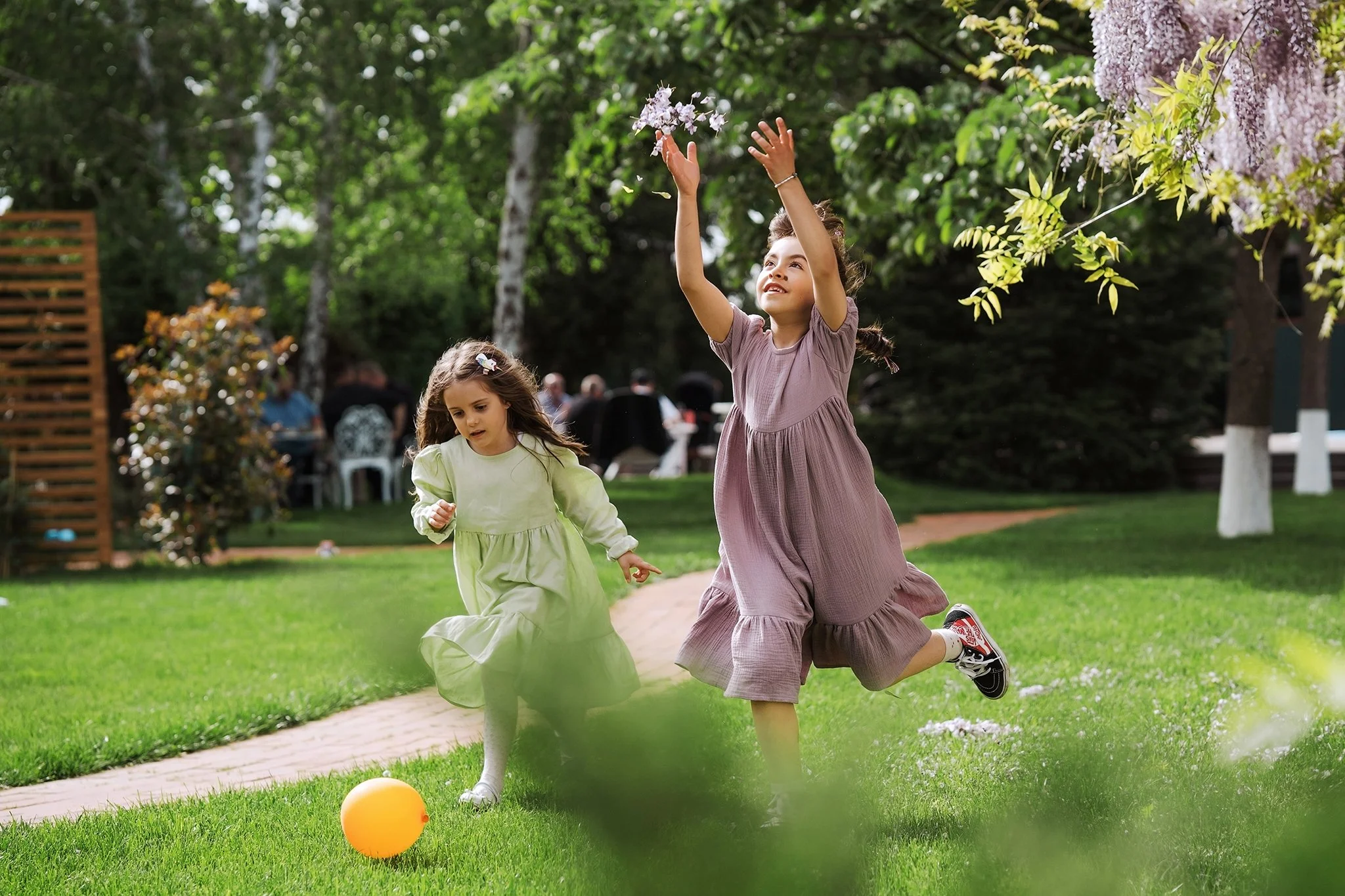 Two young girls playing with a yellow ball outdoors on a grassy lawn, surrounded by trees and other people in the background.
