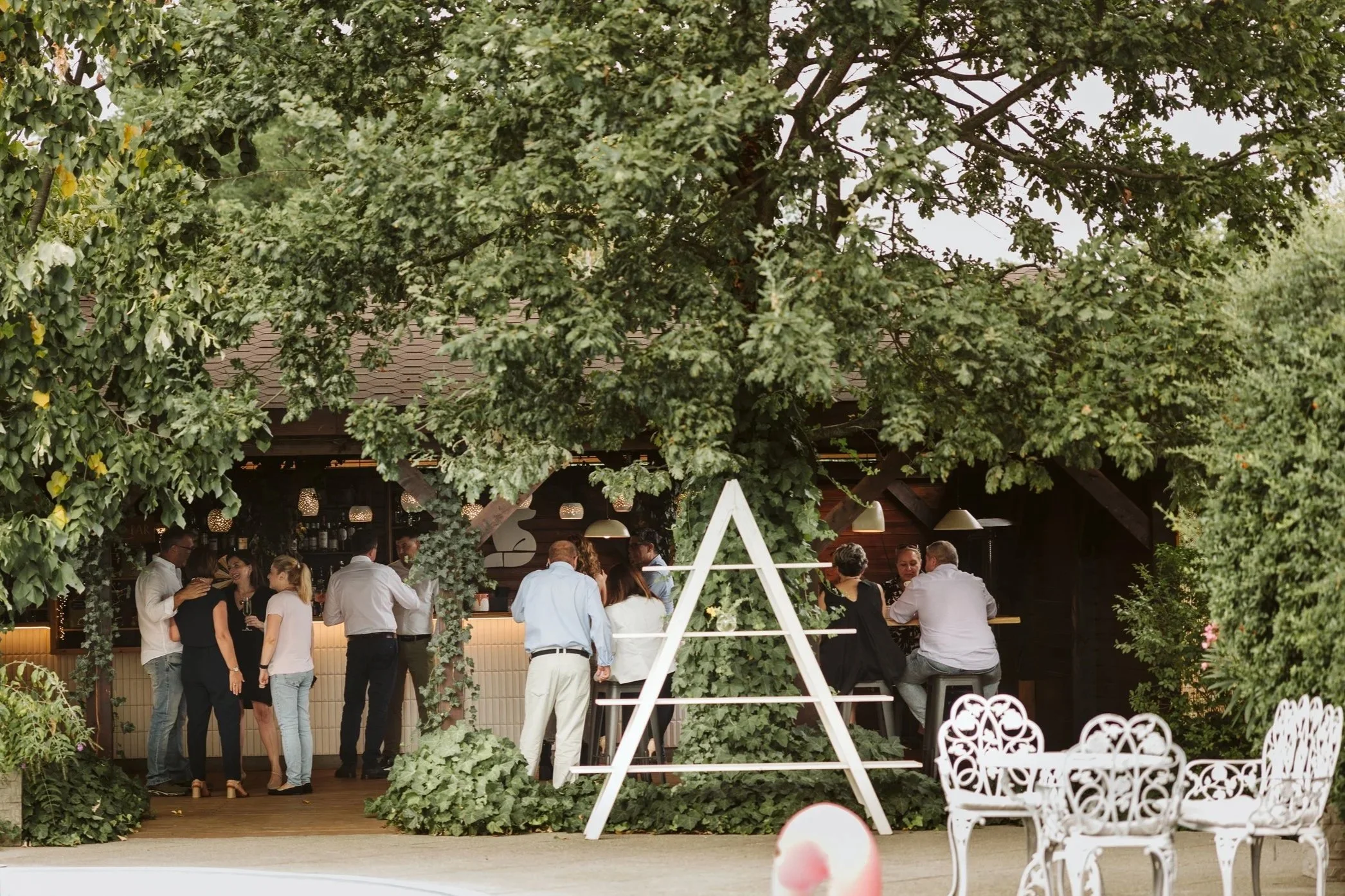 People socializing at an outdoor bar or lounge area near a swimming pool, with lush green trees and white garden furniture.