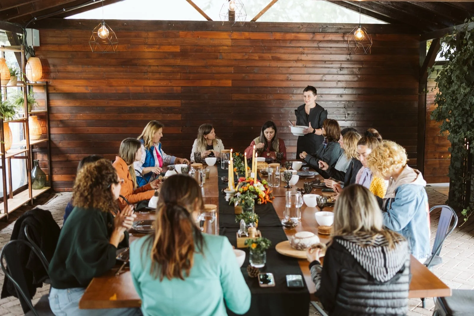 A group of people sitting around a long dining table in a rustic setting, with a woman standing at the head of the table speaking. The table is decorated with candles, flowers, and food items. The room has wooden walls, shelves with plants and lamps, and pendant lights hanging from the ceiling.