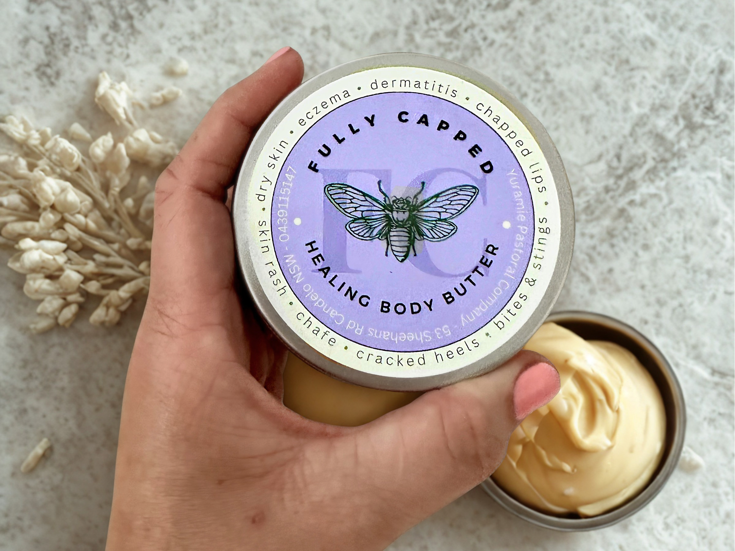 A hand holding a container of natural body butter made with beeswax with a purple and cream label featuring a bee illustration.