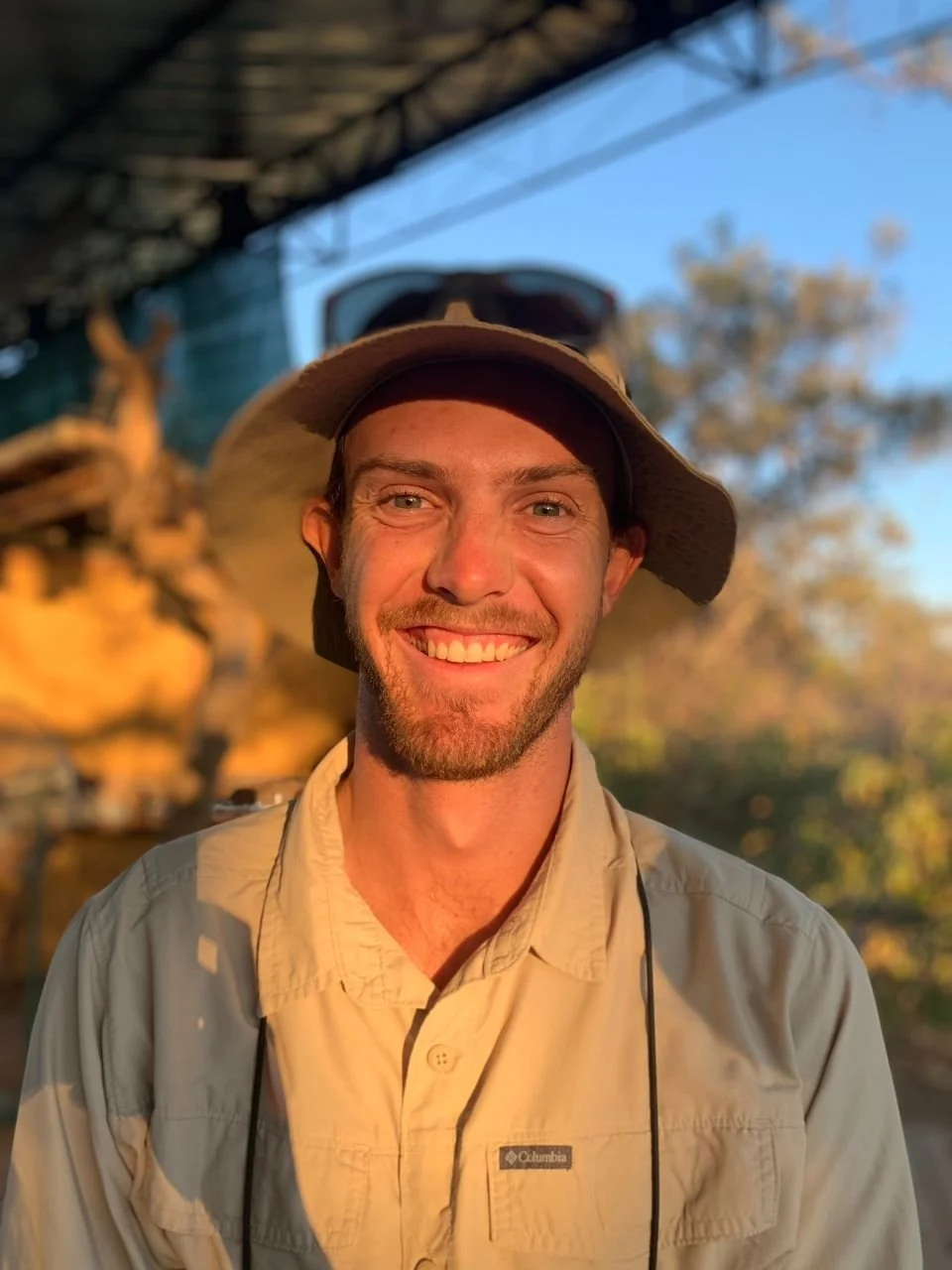 Smiling man wearing a wide-brim hat and khaki shirt outdoors with trees and blue sky in the background, sunset lighting.