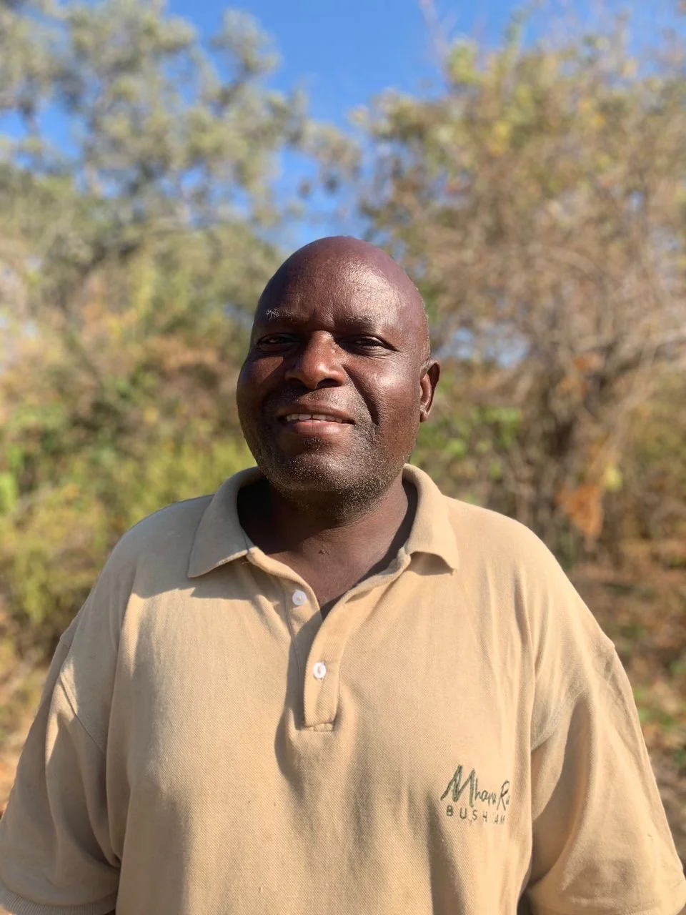 A man with dark skin, bald head, and facial hair standing outdoors with trees and blue sky in the background, wearing a beige polo shirt.