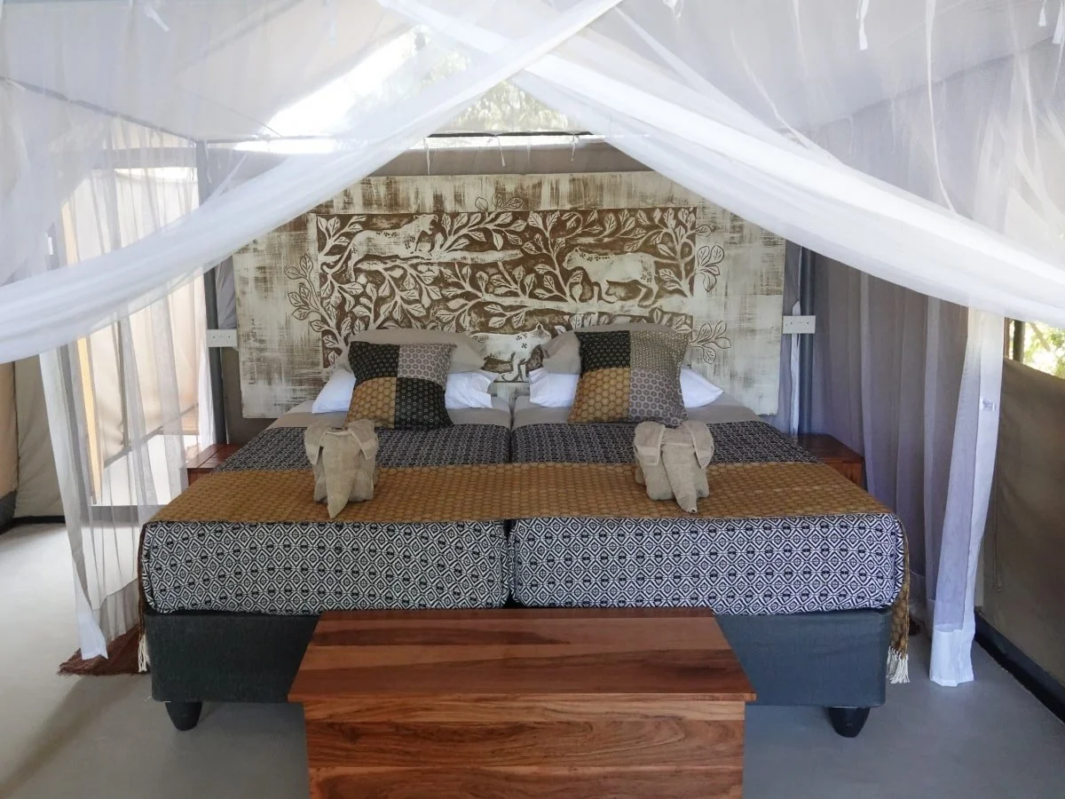 A luxurious bed inside a tent with white mosquito netting, wooden accents, and decorative pillows, with carved animal and tree artwork on the headboard.