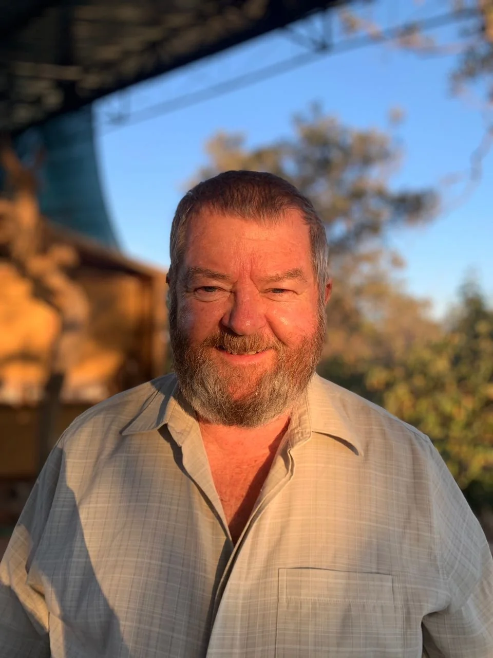A smiling middle-aged man with a beard and short hair, wearing a beige shirt, standing outdoors during sunset. There are trees and a blue sky in the background.