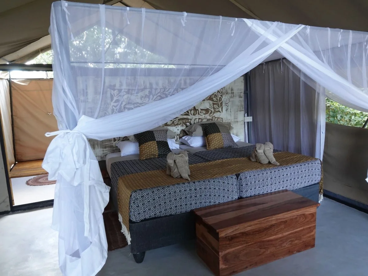 A bed with a net canopy, decorative pillows, and a wooden chest at the foot of the bed inside a tent or enclosed outdoor space.