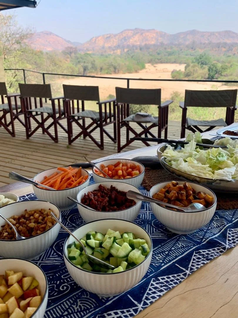 Bowls of fresh vegetables and toppings on a table outdoors, with a mountain view in the background.