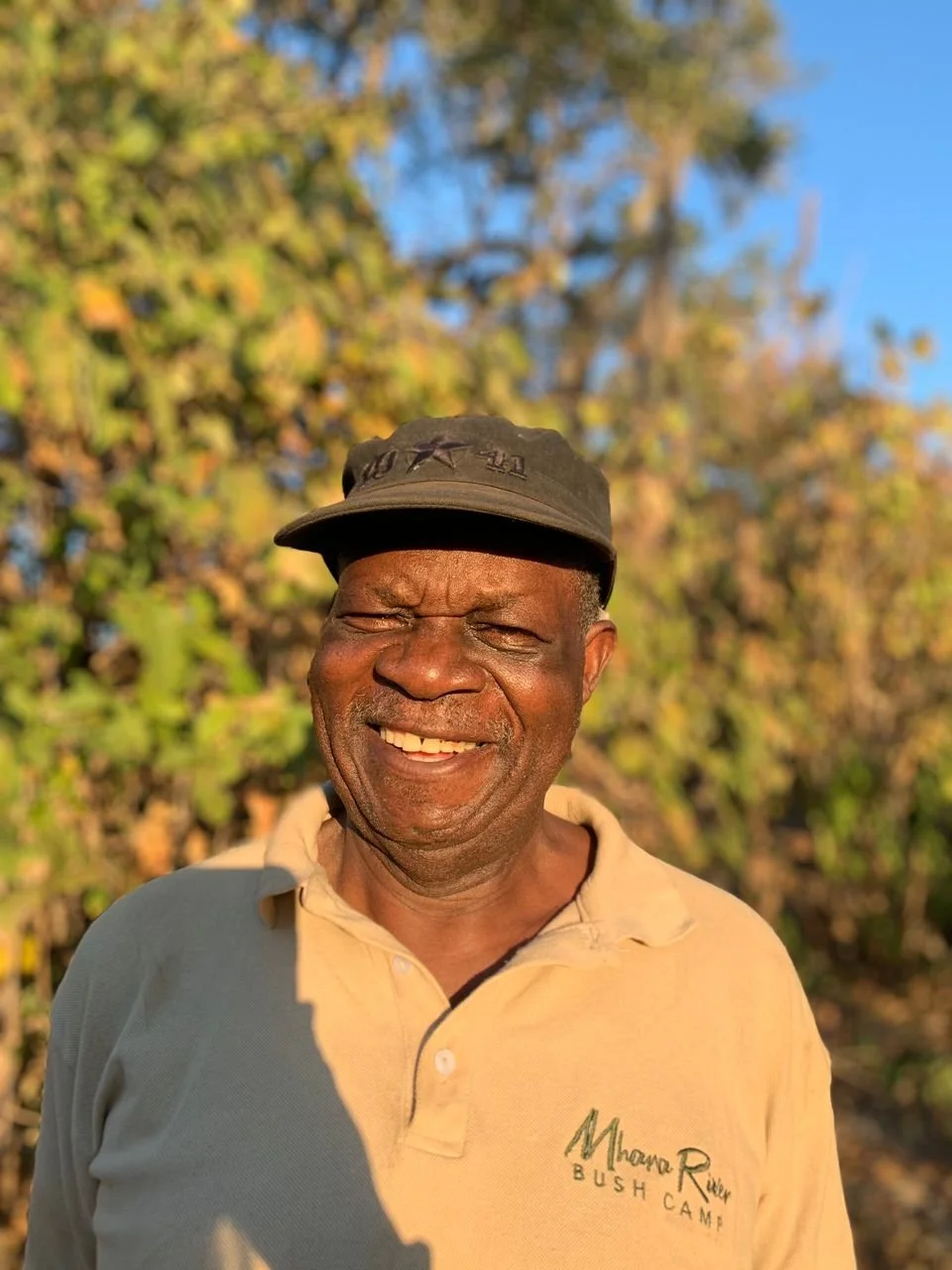 Smiling man wearing a beige polo shirt with 'Mhoroz River Bush Camp' logo and a black cap, standing outdoors with trees and blue sky in the background.