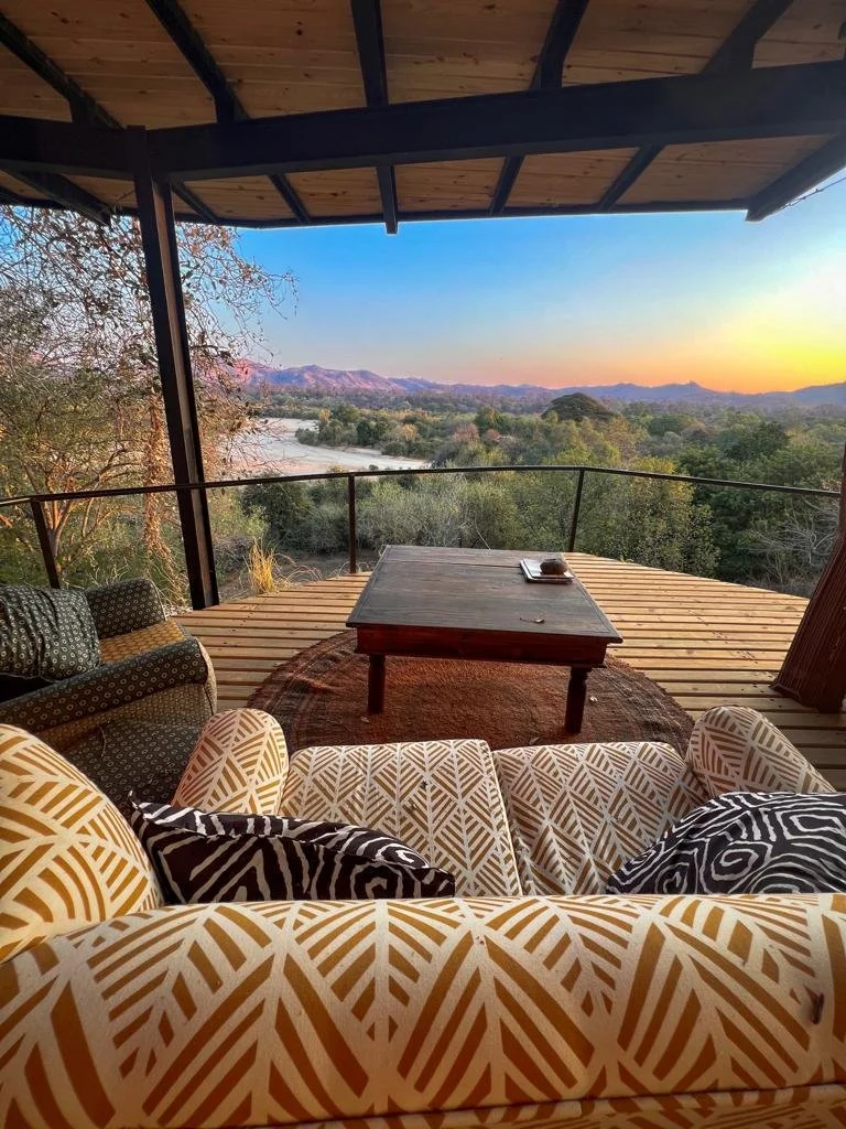 View from a cozy covered outdoor patio overlooking a river and mountains at sunset, with a wooden coffee table and patterned armchairs.