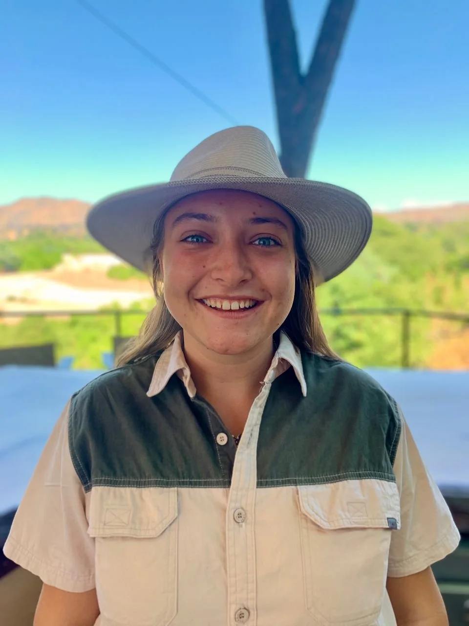 Smiling woman wearing a wide-brimmed hat and a collared shirt outdoors with hills and trees in the background.