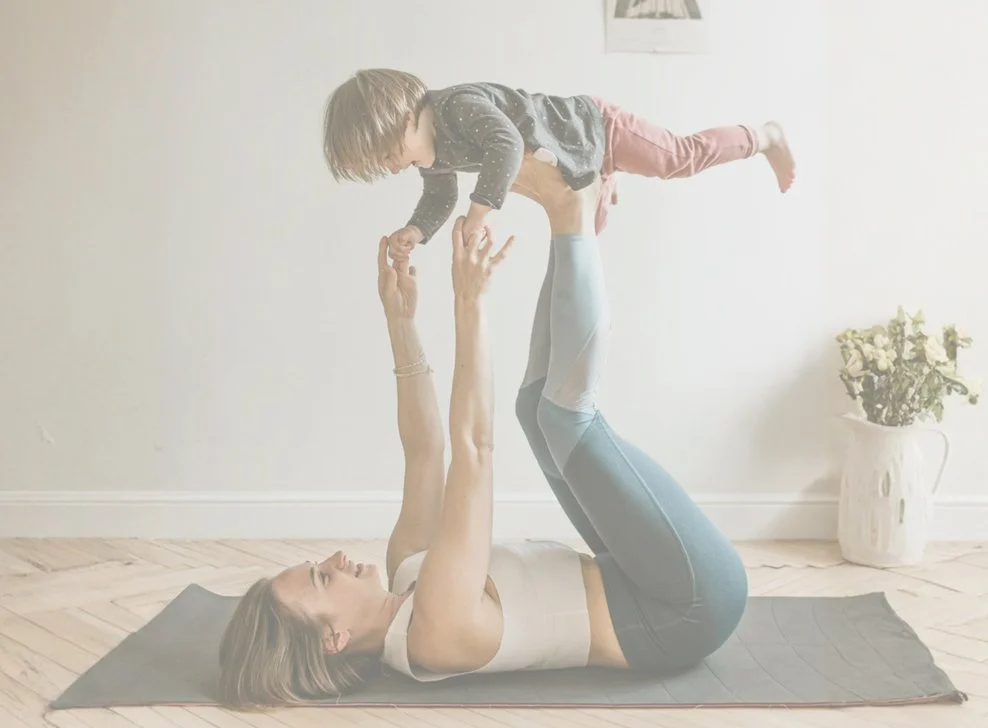 Woman lying on yoga mat lifting a young child into the air while on her back in a room with white walls and a vase of white flowers.
