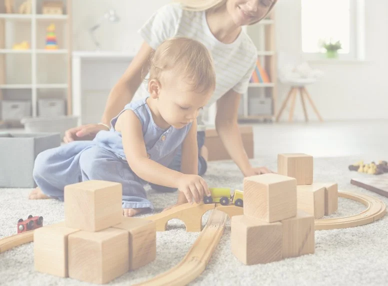 A young girl playing with wooden toy train set while an adult woman watches in a bright, modern living room with shelves and natural light.