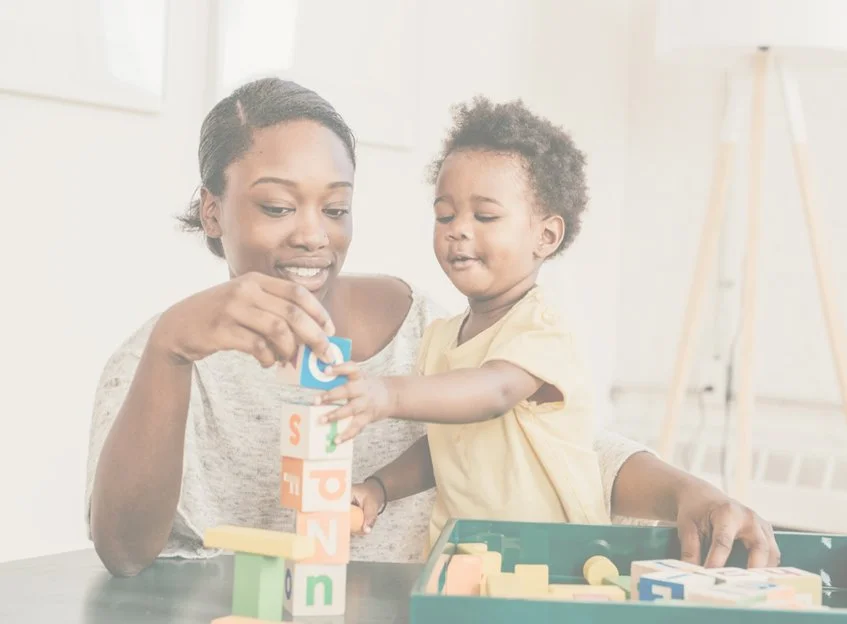 A woman and a young child playing with colorful alphabet blocks at a table, indoors.