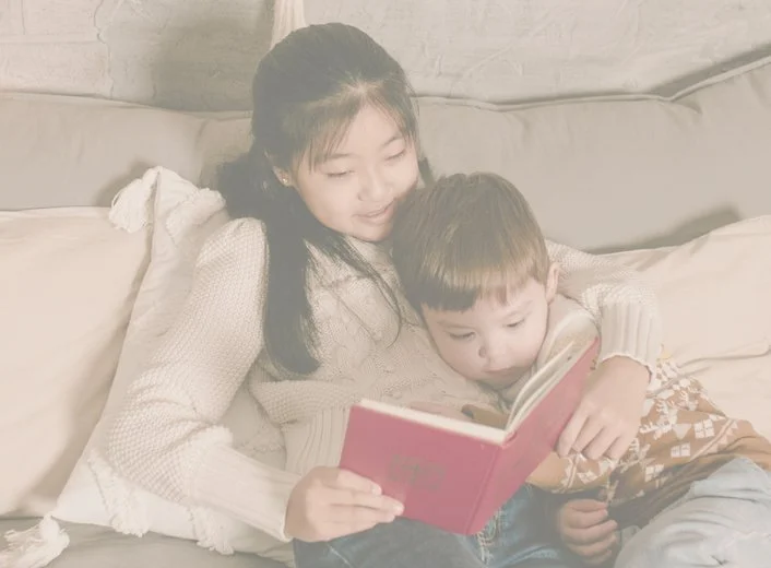 A mother and son sitting on a couch, reading a book together, with the mum smiling and the boy focused on the book.