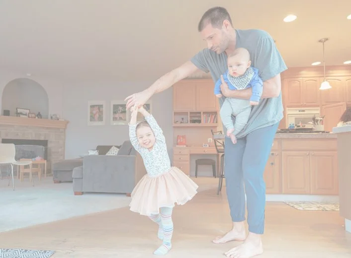 A daddy dancing with a young girl holding hands, while holding a baby in his other arm inside a cozy, well-lit home.
