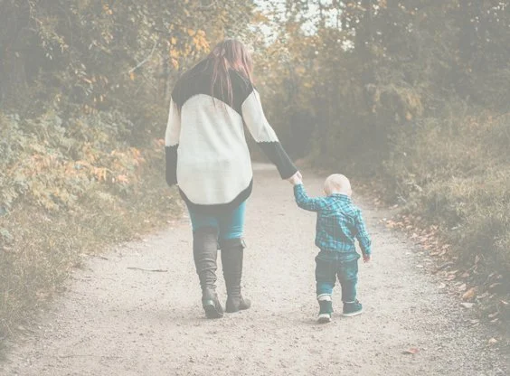 A woman holding the hand of a young child as they walk down a dirt path surrounded by trees.