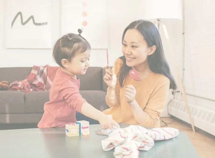 A woman and a young girl playing with toys at a table in a bright living room.