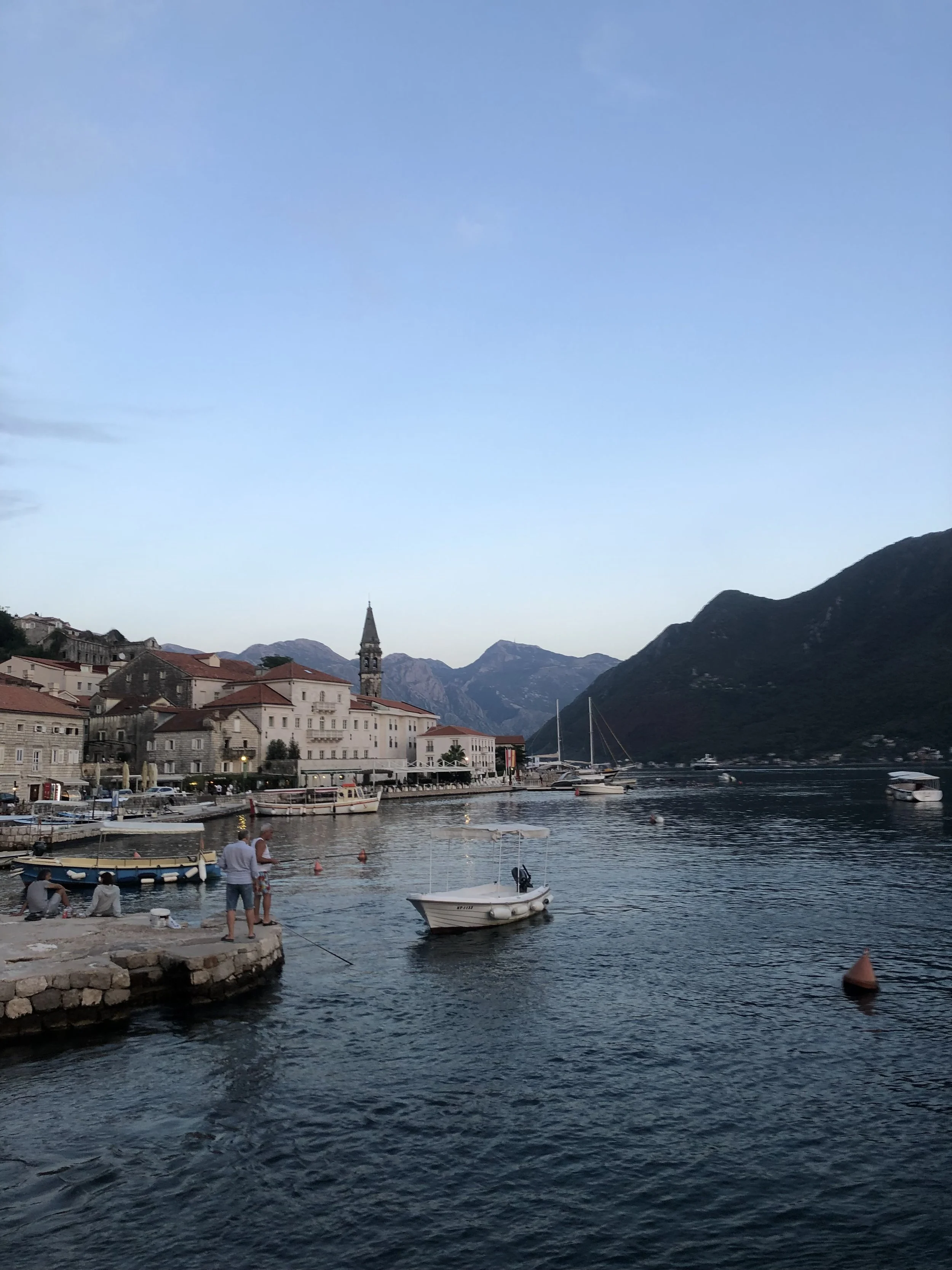 A scenic harbor in with boats docked at Perast, Montenegro. Mountain range in the background, and people enjoying the waterfront.
