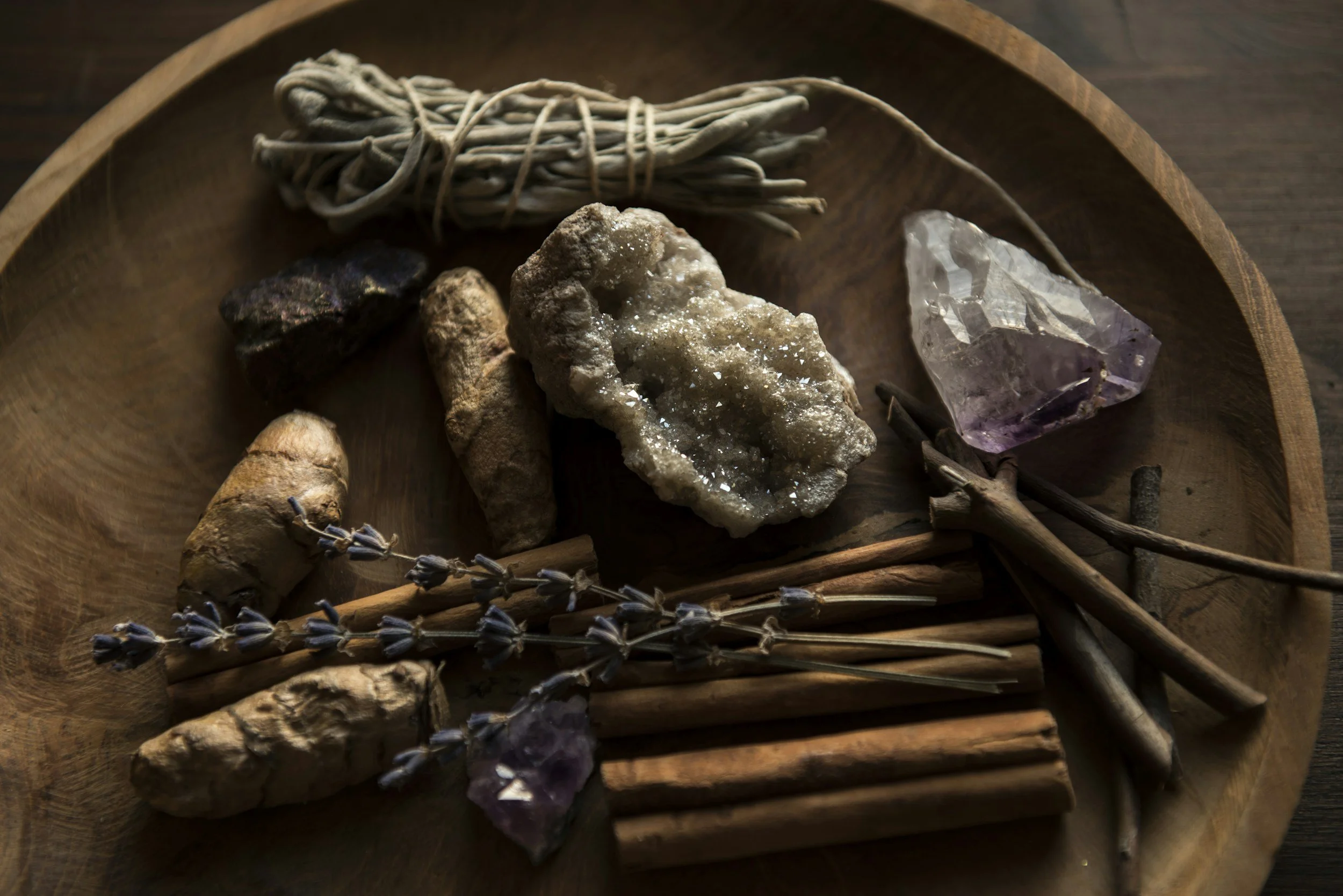 Various natural crystals, dried lavender, cinnamon sticks, ginger root, and sage bundles arranged on a wooden tray.