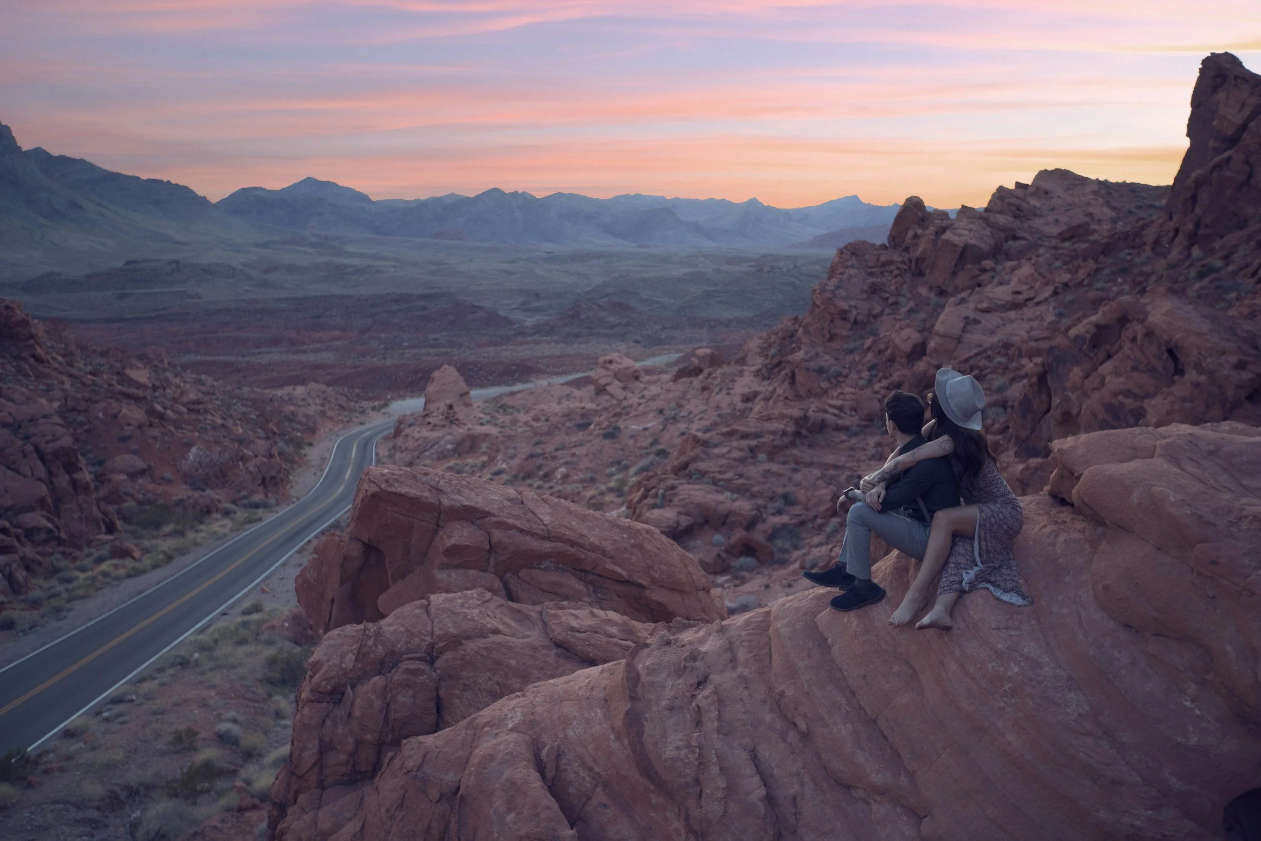 A couple sitting on red rocky desert terrain overlooking a winding road and mountain landscape at sunset.
