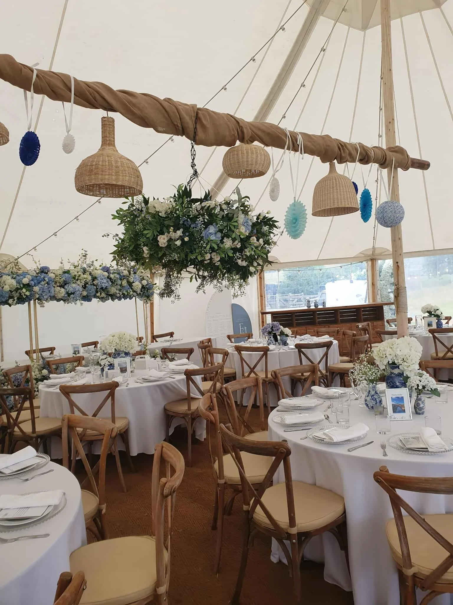 Interior of a decorated event tent with round tables covered in white tablecloths, set with plates and cutlery, surrounded by wooden chairs. Hanging above are woven lamps and decorative hanging spheres, with a large floral arrangement of greenery and white and blue flowers suspended mid-air.