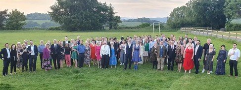 A large group of people standing outdoors on a grassy field with trees and hills in the background.