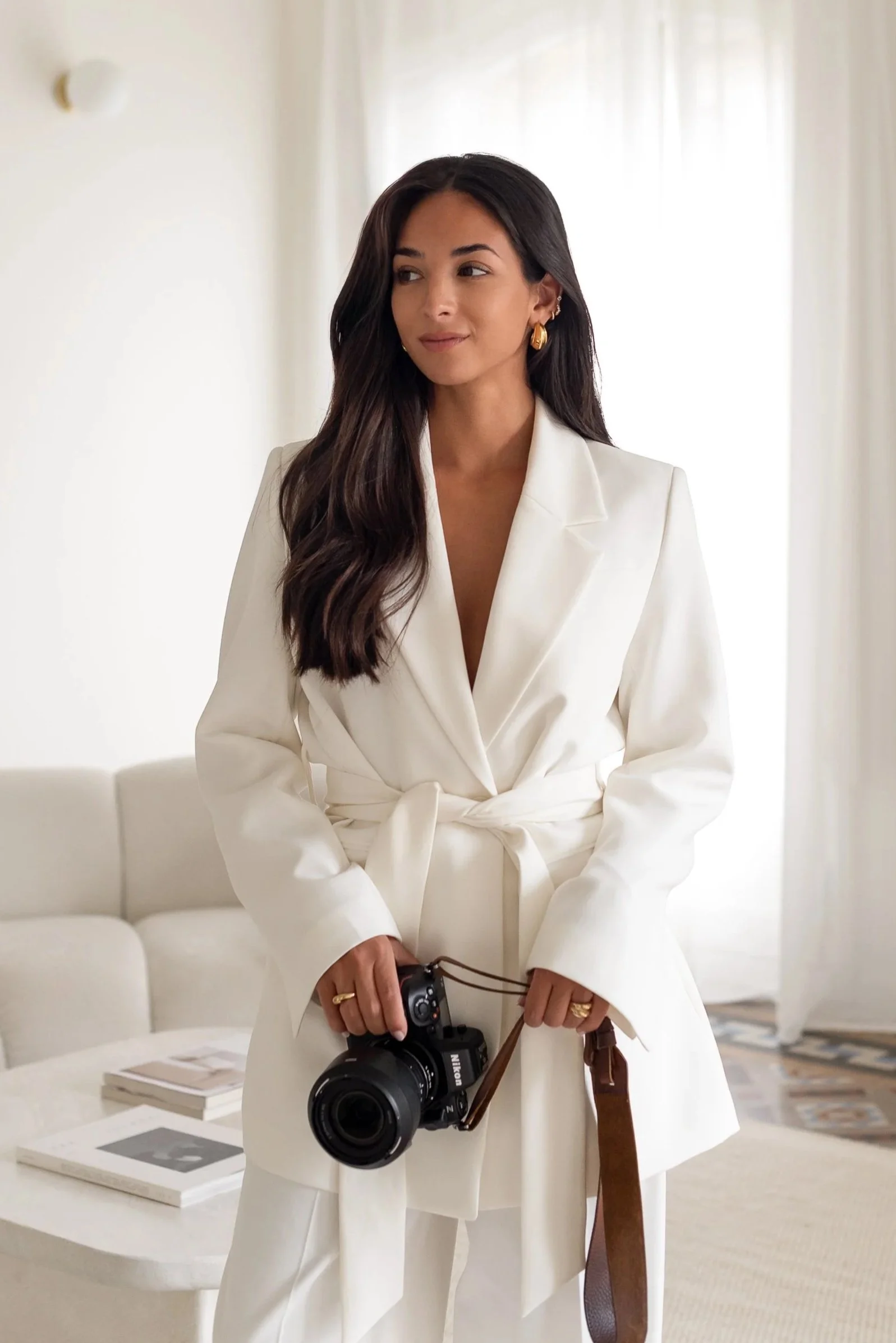 A woman with long dark hair, wearing a white blazer and pants, holding a Nikon camera, standing in a bright room with white walls and curtains, a white sofa, magazines, and books on a coffee table in the background.
