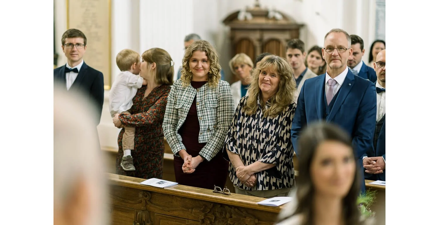 Gäste und Familie während der kirchlichen Trauung im Kloster Irsee, aufmerksam und emotional in den Kirchenbänken stehend, während der Zeremonie.