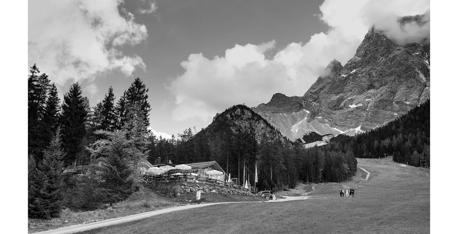Weitläufige Berglandschaft bei Ehrwald mit Blick auf die Zugspitze, Gamsalm am Hang und kleine Hochzeitsgesellschaft auf einer Wiese in Schwarzweiß
