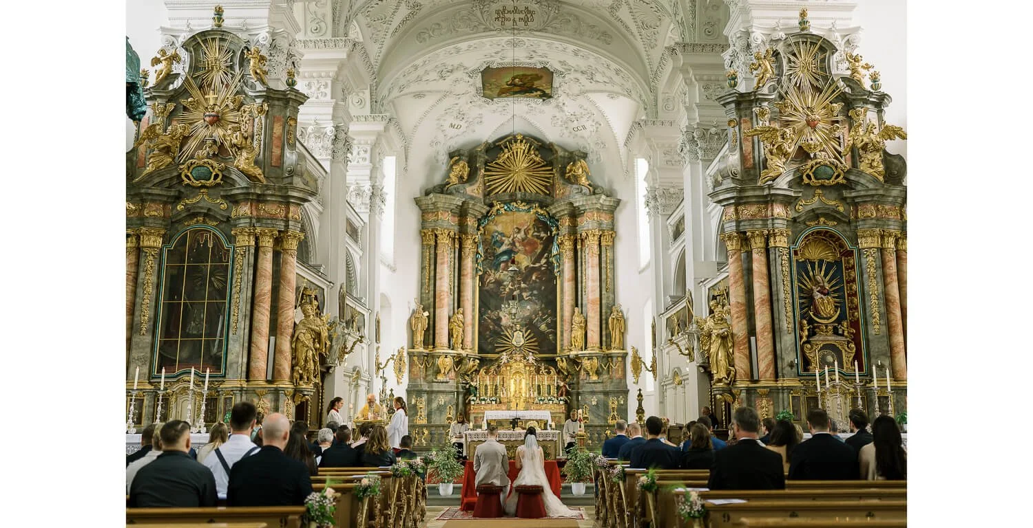 Kirchliche Trauung im Kloster Irsee, Braut und Bräutigam knien vor dem Altar, Gäste sitzen in den Kirchenbänken, barocker Innenraum mit reich verziertem Hochaltar.