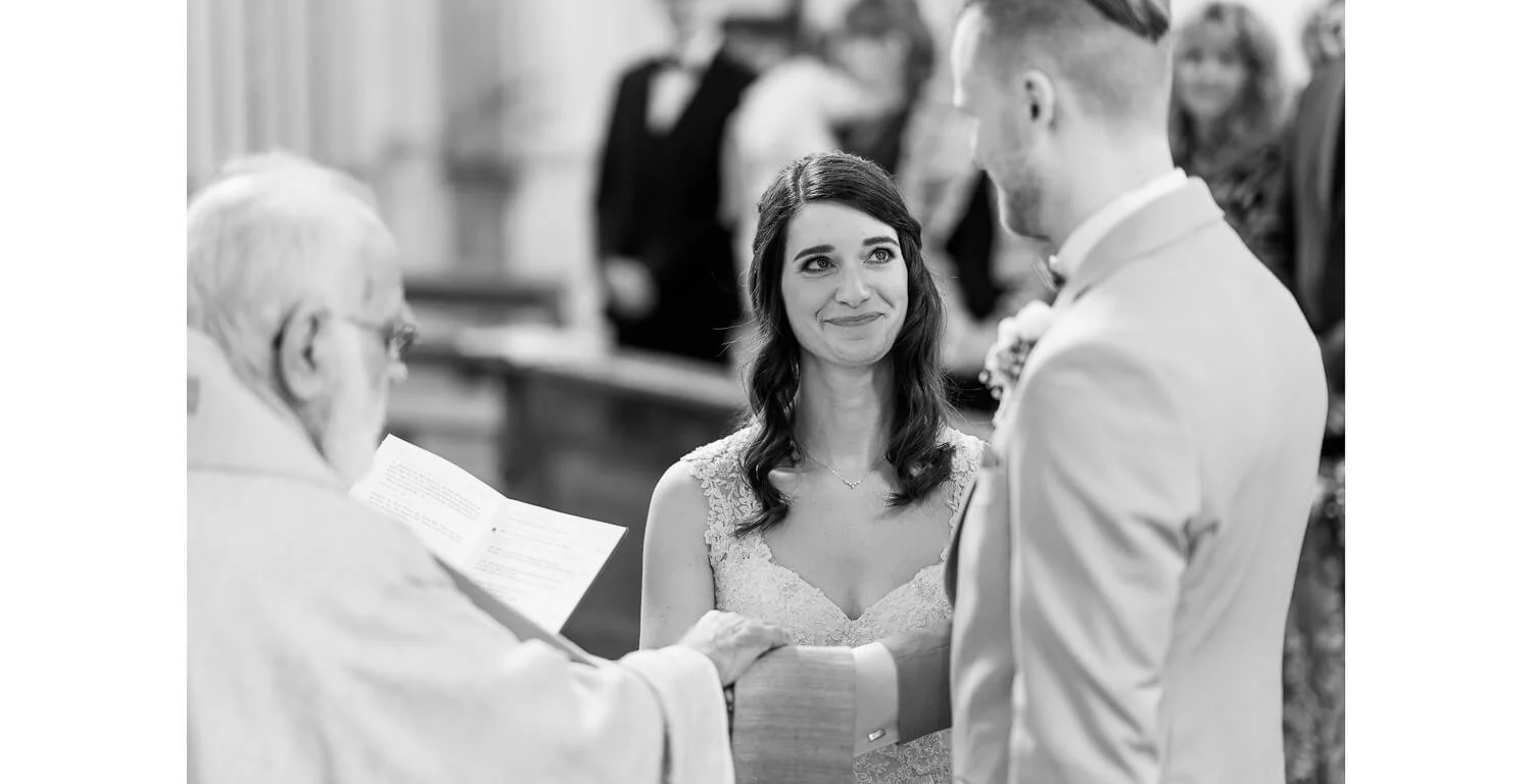 Schwarz-weiß Aufnahme während der kirchlichen Trauung im Kloster Irsee: Braut und Bräutigam stehen sich gegenüber, halten sich an den Händen, während der Priester den Trauspruch vorliest.