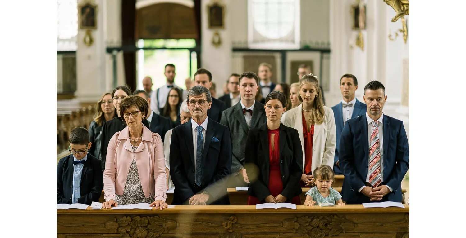 Hochzeitsgäste stehen während der kirchlichen Trauung im Kloster Irsee in den Kirchenbänken und verfolgen die Zeremonie aufmerksam