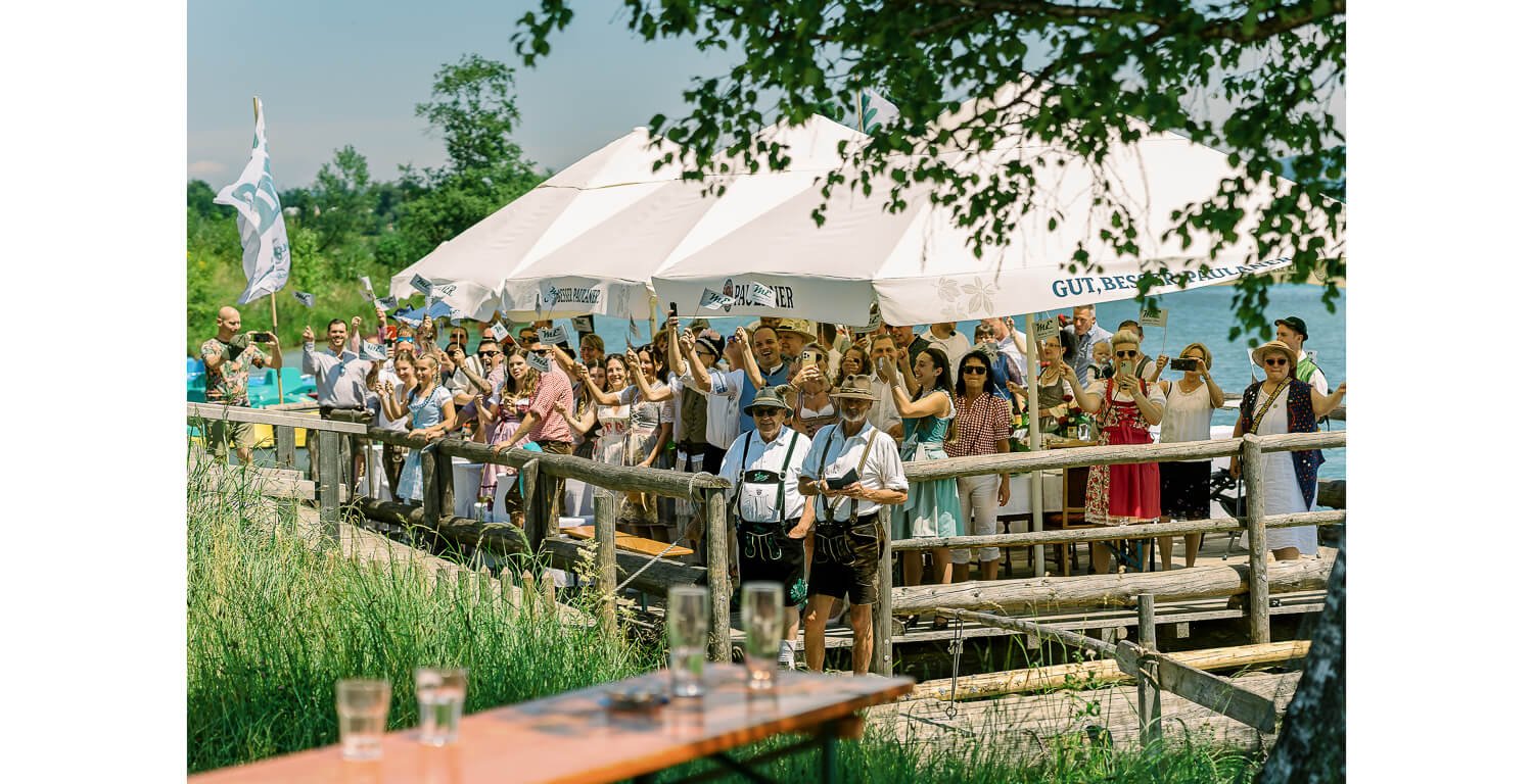 Gäste in traditioneller Tracht stehen am Ufer in Lechbruck am See und begrüßen das Brautpaar bei einer standesamtlichen Hochzeit auf einem Floß