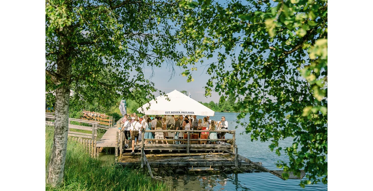 Gäste bei der standesamtlichen Hochzeit auf dem Floß in Lechbruck am See, versammelt unter Sonnenschirmen am Ufer