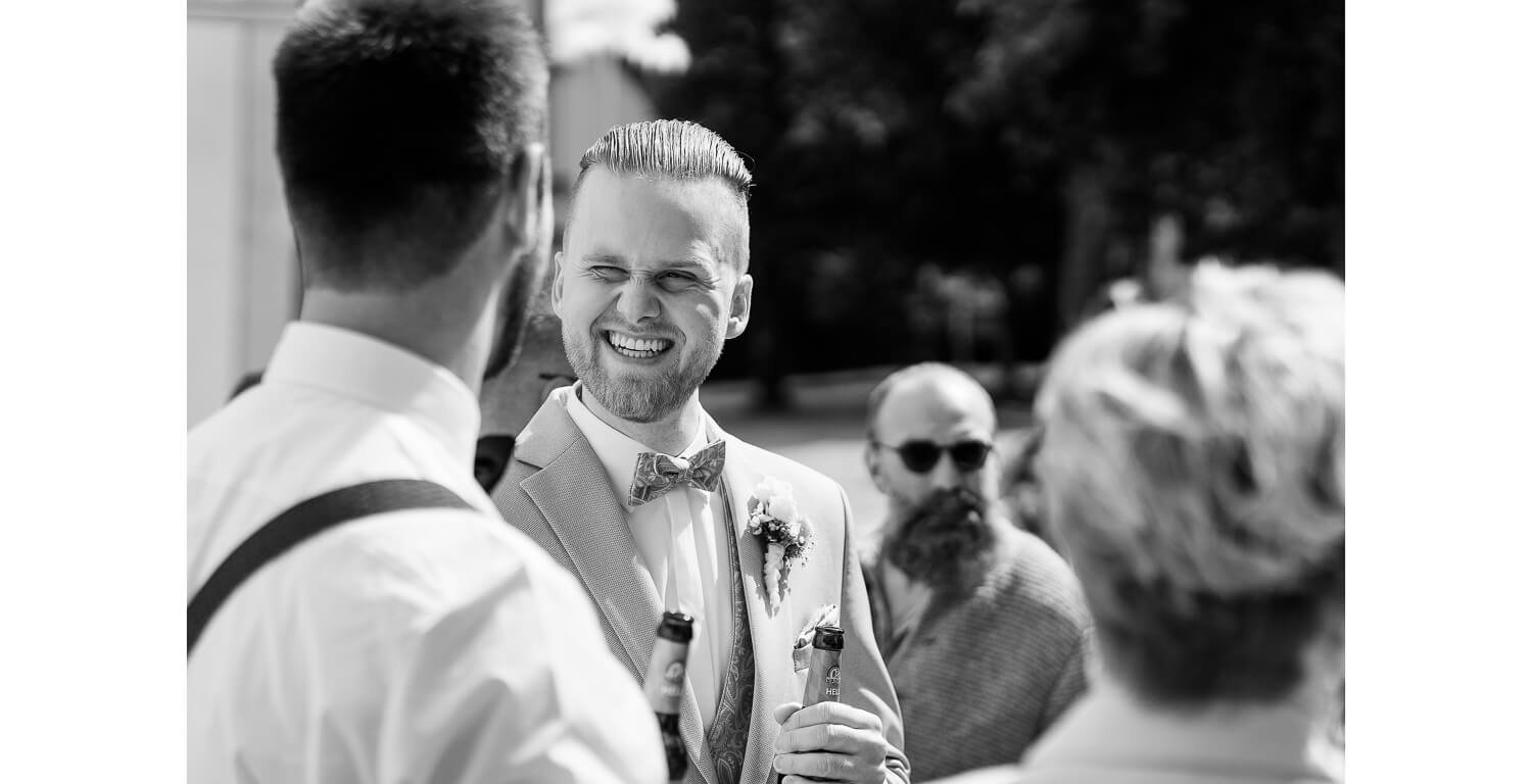 Bräutigam lacht nach der kirchlichen Trauung im Kloster Irsee im Gespräch mit Hochzeitsgästen im Außenbereich